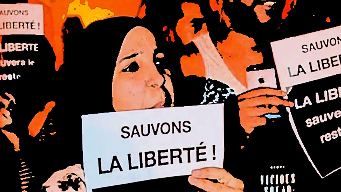 The image depicts a group of people holding up signs with messages in French. The main focus is on two individuals in the foreground. One is wearing a hijab and both are raising their signs high, which read "Sauvons la liberté!" (Save Liberty!). The atmosphere suggests a protest or rally, with a sense of solidarity and determination among the participants. The background is filled with more people and signs, contributing to the overall theme of advocating for freedom.