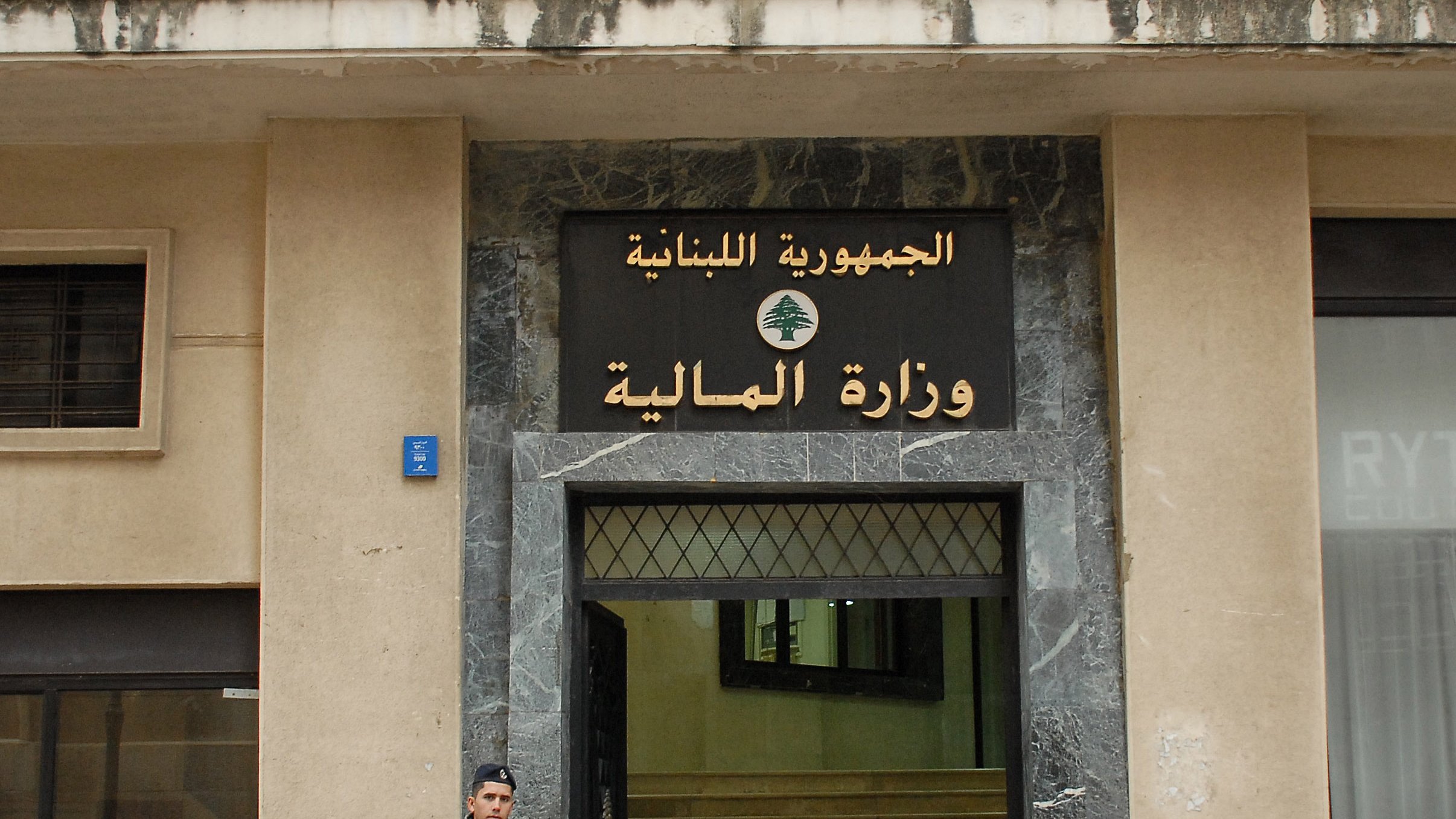 الصورة تظهر واجهة وزارة المالية في لبنان، والتي تحمل العلامة الرسمية للجمهورية اللبنانية. يوجد على الجهة اليسرى من الصورة جندي يقف حارسًا، يحمل سلاحًا، وقد ارتدى زيًا عسكريًا. المبنى يبدو بسيطًا وعصريًا، ويعكس الطراز المعماري الحضري.