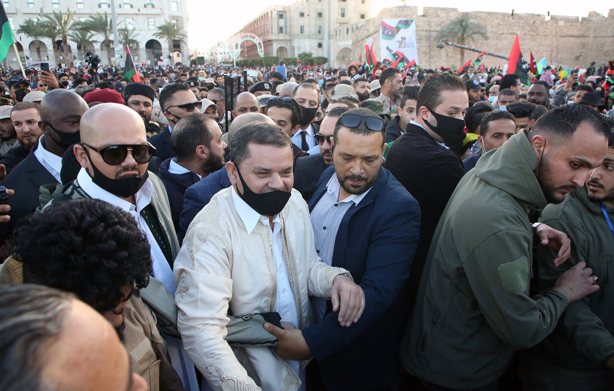 الصورة تُظهر حشودًا كبيرة من الناس في مكان عام، بعضهم يرتدي الكمامات، ويبدو أن هناك شخصيات بارزة في الوسط. يظهر الأشخاص في الصورة ممالأة وتفاعل مع بعضهم، مما يوحي بأن هناك حدثًا مهمًا يجري. يمكن أن يكون الحدث مظاهرة أو تجمعًا سياسيًا، حيث تحمل الأعلام وتظهر بعض الوجوه المألوفة. الجو بشكل عام يبدو متوترًا ولكن مليئًا بالحماس.