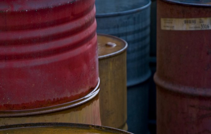 L'image montre plusieurs fûts en métal empilés les uns sur les autres. Chaque fût a une couleur différente : rouge, vert, noir et jaune. La surface des fûts semble briller, probablement à cause d'un liquide qui s'y trouve, reflétant la lumière. L'ensemble de la composition donne une impression de dépôt industriel, avec une texture rustique et des nuances de couleurs intéressantes.