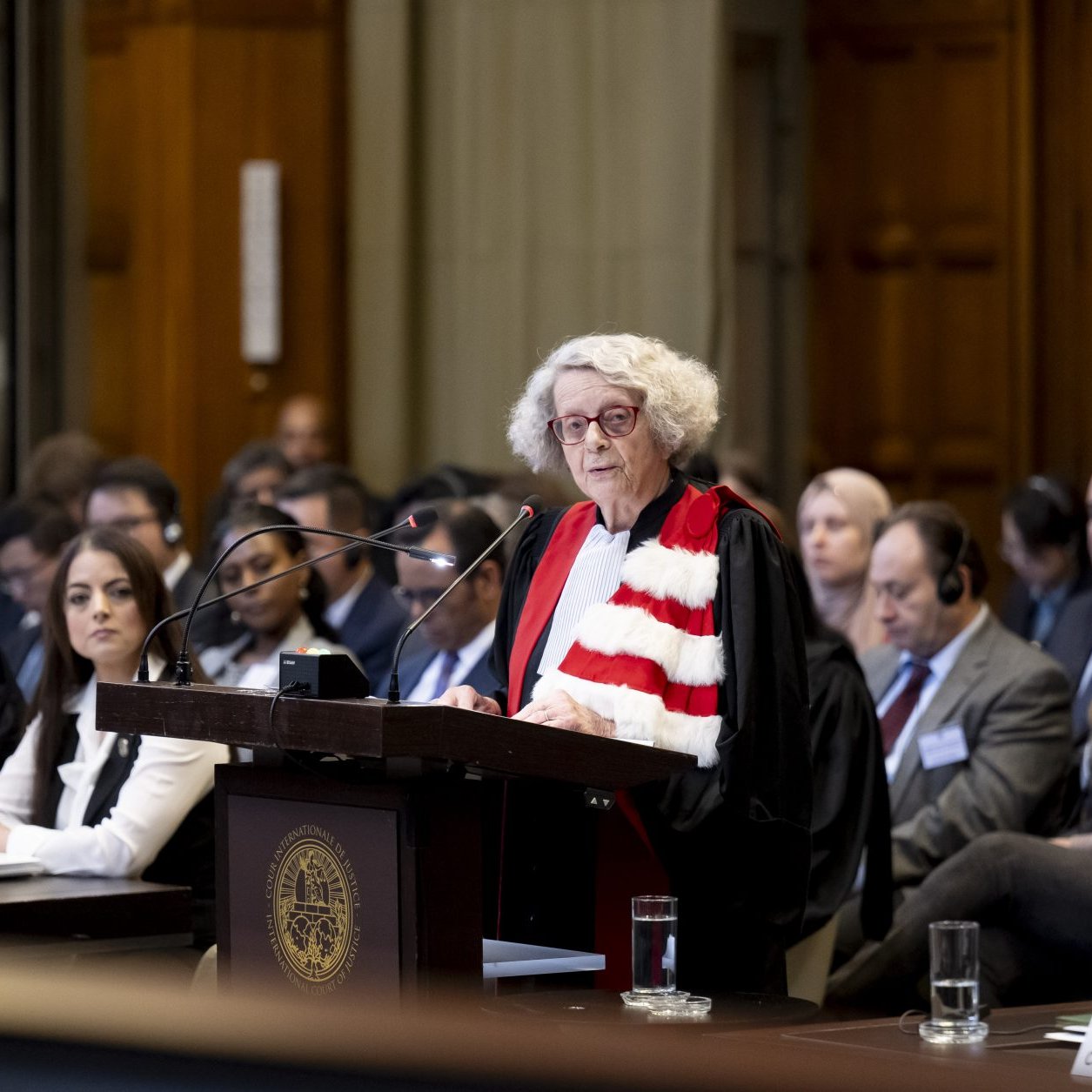 L'image montre une scène dans une salle d'audience, probablement dans un tribunal. Une femme, vêtue d'une robe magistrale et d'une écharpe, se tient derrière un pupitre en train de prendre la parole. Plusieurs personnes sont assises à des tables devant elle, écoutant attentivement. L'environnement semble formel, avec des éléments architecturaux en bois et des fenêtres qui apportent de la lumière naturelle. L'atmosphère est solennelle et concentrée, indicative d'une procédure judiciaire importante.