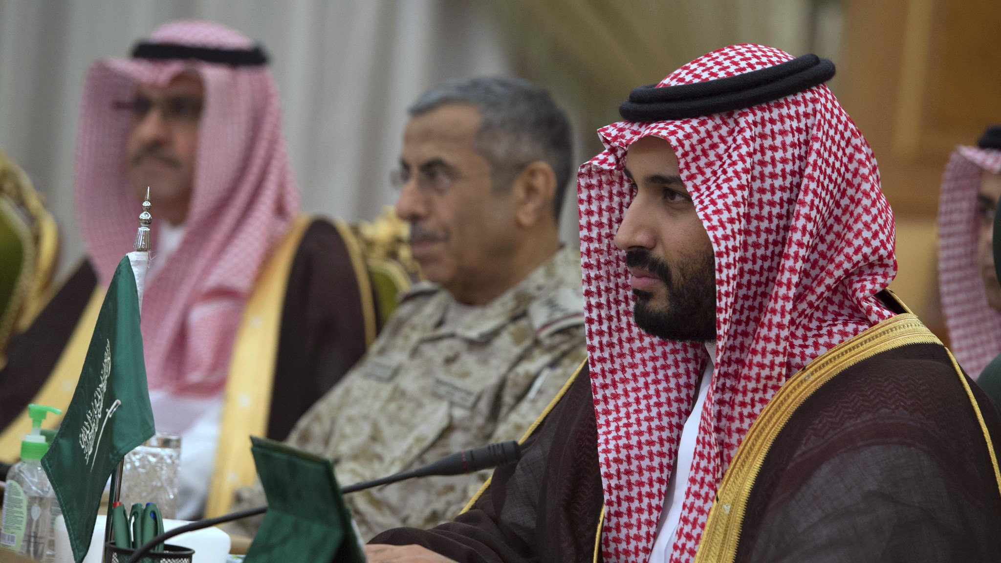 The image shows a group of individuals in formal attire, seated around a table. The man in the foreground is dressed in traditional Saudi attire, wearing a red and white checkered headscarf (ghutrah) and a long brown robe. He appears focused, looking ahead. In the background, another man in military attire is also present, along with other officials. The setting appears to be a formal meeting or conference, indicated by the presence of flags and documents on the table.