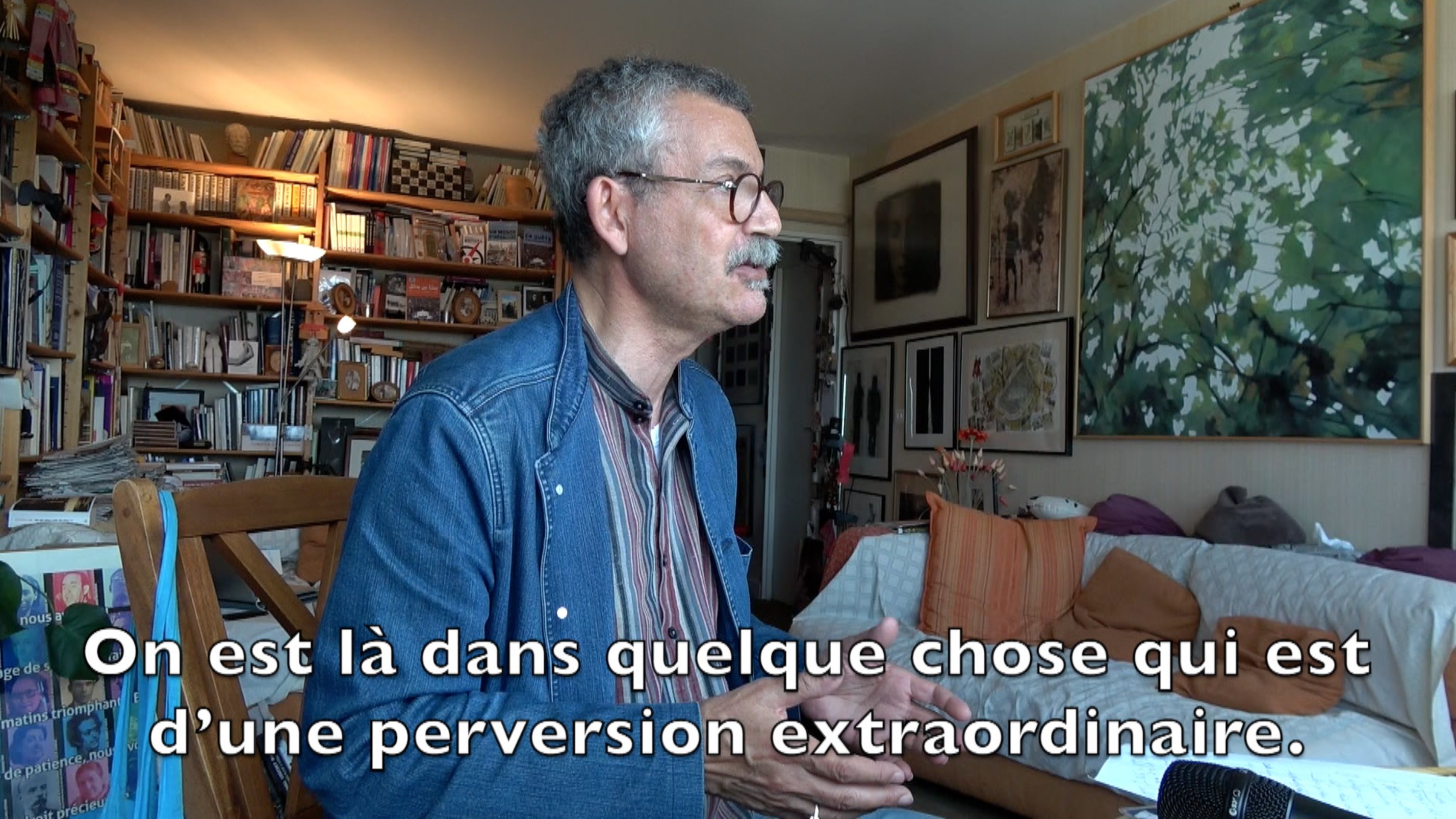 L'image montre un homme assis à une table, parlant avec animation. Il semble s'exprimer sur un sujet profond, probablement lié à des thèmes intellectuels ou sociaux. En arrière-plan, on peut voir des étagères remplies de livres et plusieurs œuvres d'art accrochées aux murs, ce qui indique un environnement artistique et studieux. Le texte sur l'image suggère qu'il aborde des concepts de manière critique, impliquant une réflexion sur des questions de moralité ou de société.