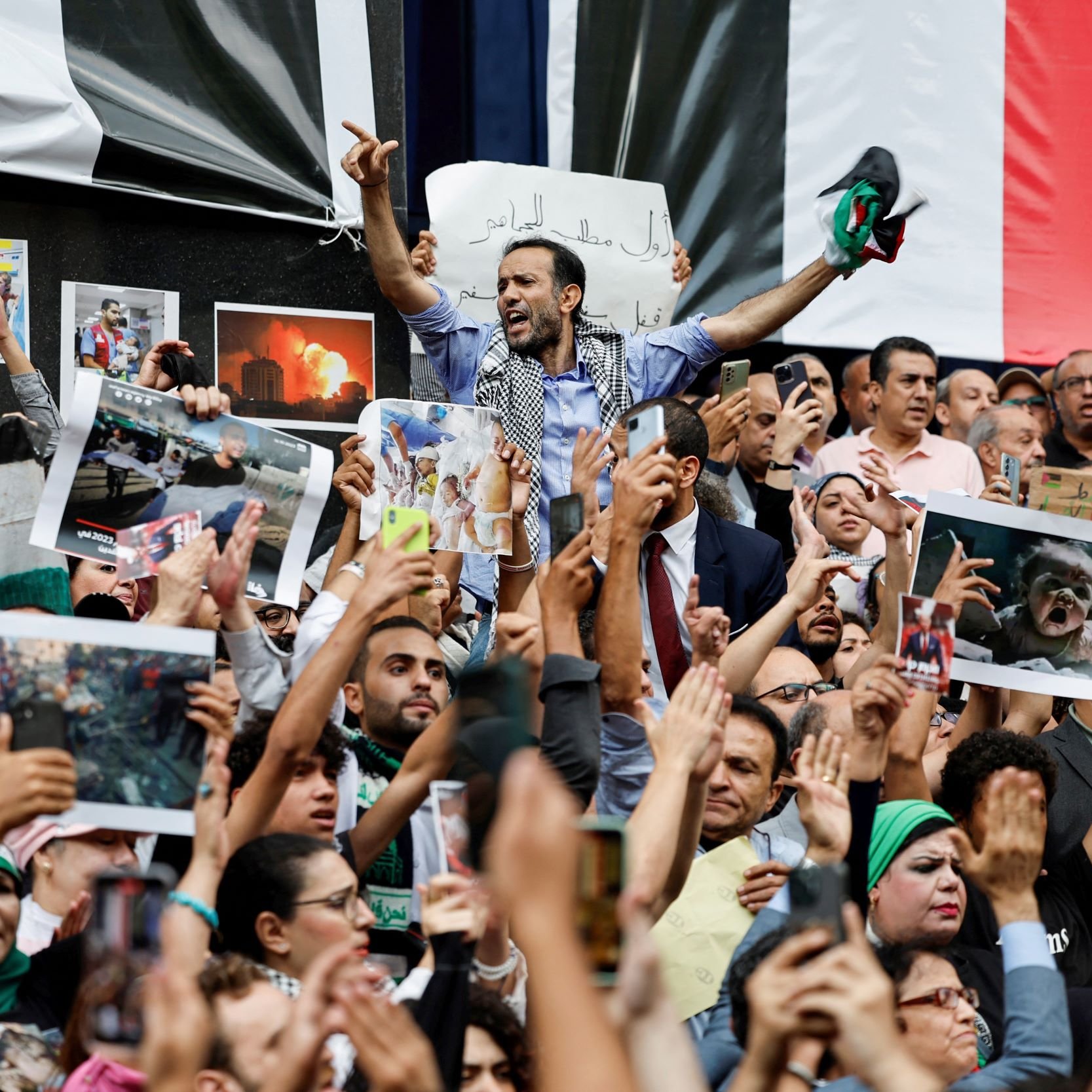 L'immagine mostra una folla di persone radunate in una manifestazione. Gli individui alzano cartelli e fotografie, esprimendo chiaramente le loro emozioni e il loro supporto per una causa. Alcuni di loro indossano sciarpe e altri simboli, mentre l'atmosfera sembra essere intensa e piena di passione. Sullo sfondo, ci sono bandiere che suggeriscono un contesto politico o sociale significativo.