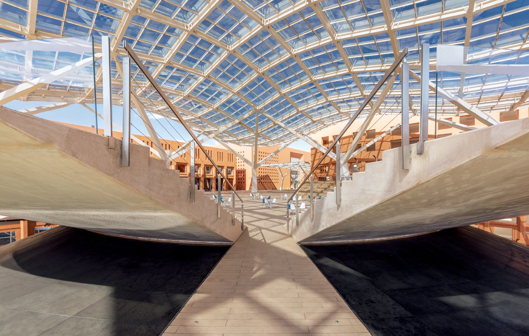 تظهر الصورة تصميمًا معماريًا حديثًا يتميز بسقف زجاجي ضخم يوفر إضاءة طبيعية وفيرة. الأرضية تحت السقف موزعة بطريقة فريدة، حيث تبرز ممرات خشبية وسطحيات مستوية، وتتنوع الأشكال الهندسية بين المنحنيات والزوايا الحادة. يعتبر هذا التصميم تجسيدًا للتوازن بين الطبيعة والتكنولوجيا، مما يعكس أسلوبًا عصريًا ومبتكرًا في البناء.