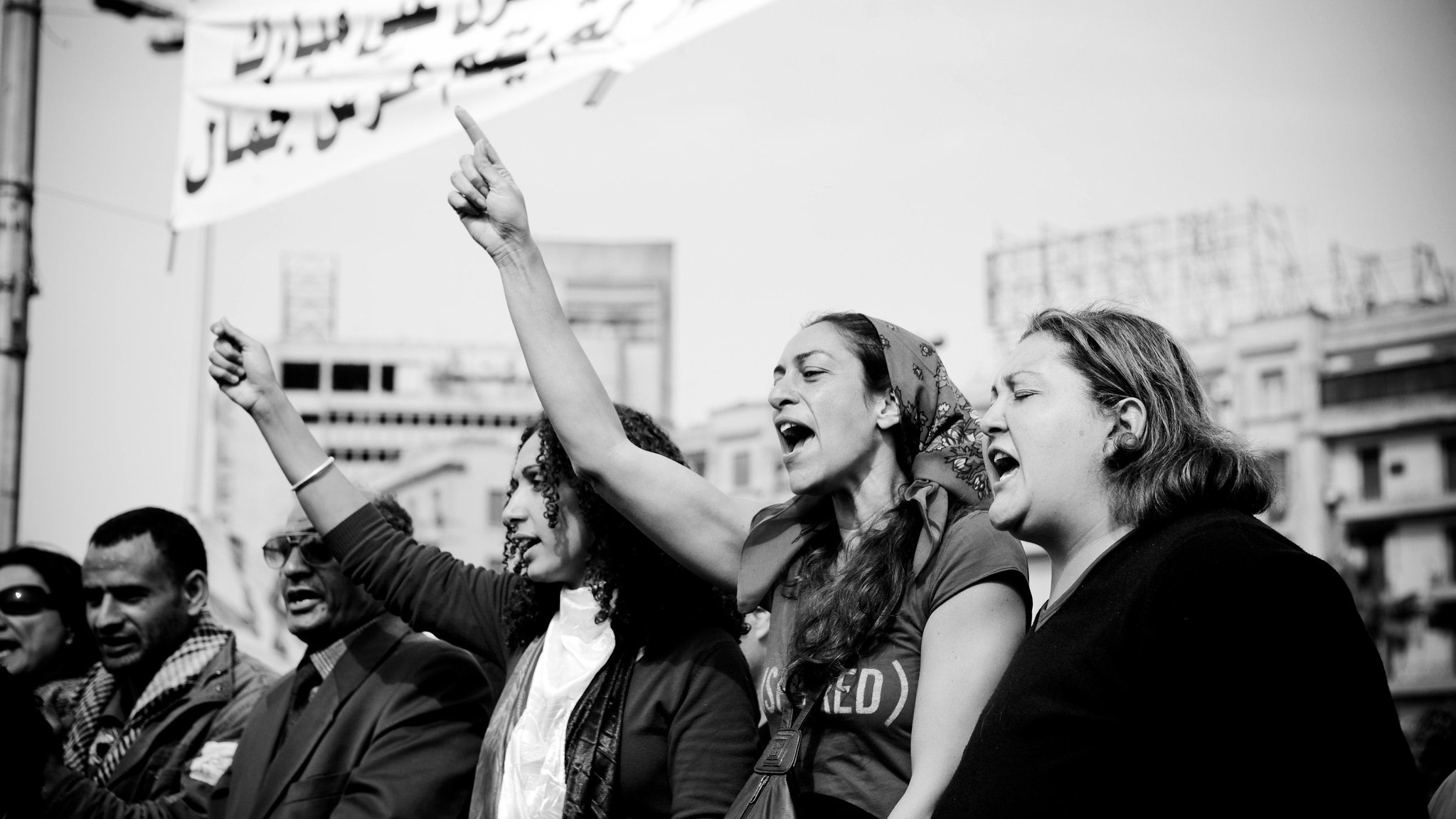 L'image montre un groupe de personnes participant à une manifestation. On peut voir des femmes au premier plan levant le poing et chantant, exprimant des émotions fortes et de la détermination. L'ambiance semble dynamique et engageante. En arrière-plan, des banderoles apparaissent, suggérant un message ou une demande collective. La photo est en noir et blanc, ce qui accentue l'intensité et l'impact visuel de la scène.