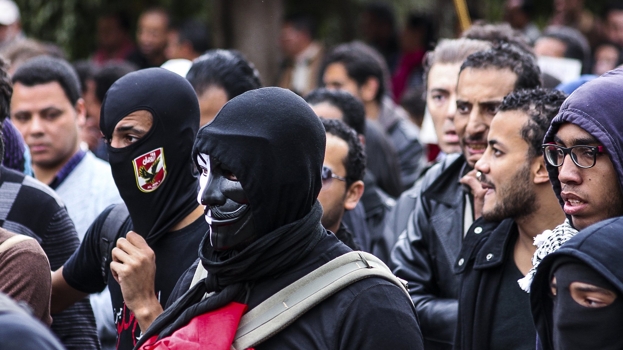 L'image montre un groupe de personnes rassemblées, probablement lors d'une manifestation. Certains portent des masques noirs, tandis que d'autres affichent des expressions déterminées. On peut observer un mélange de vêtements allant des vestes en cuir à des écharpes. L'ambiance semble être à la fois sérieuse et engagée, avec des signes de solidarité entre les participants.