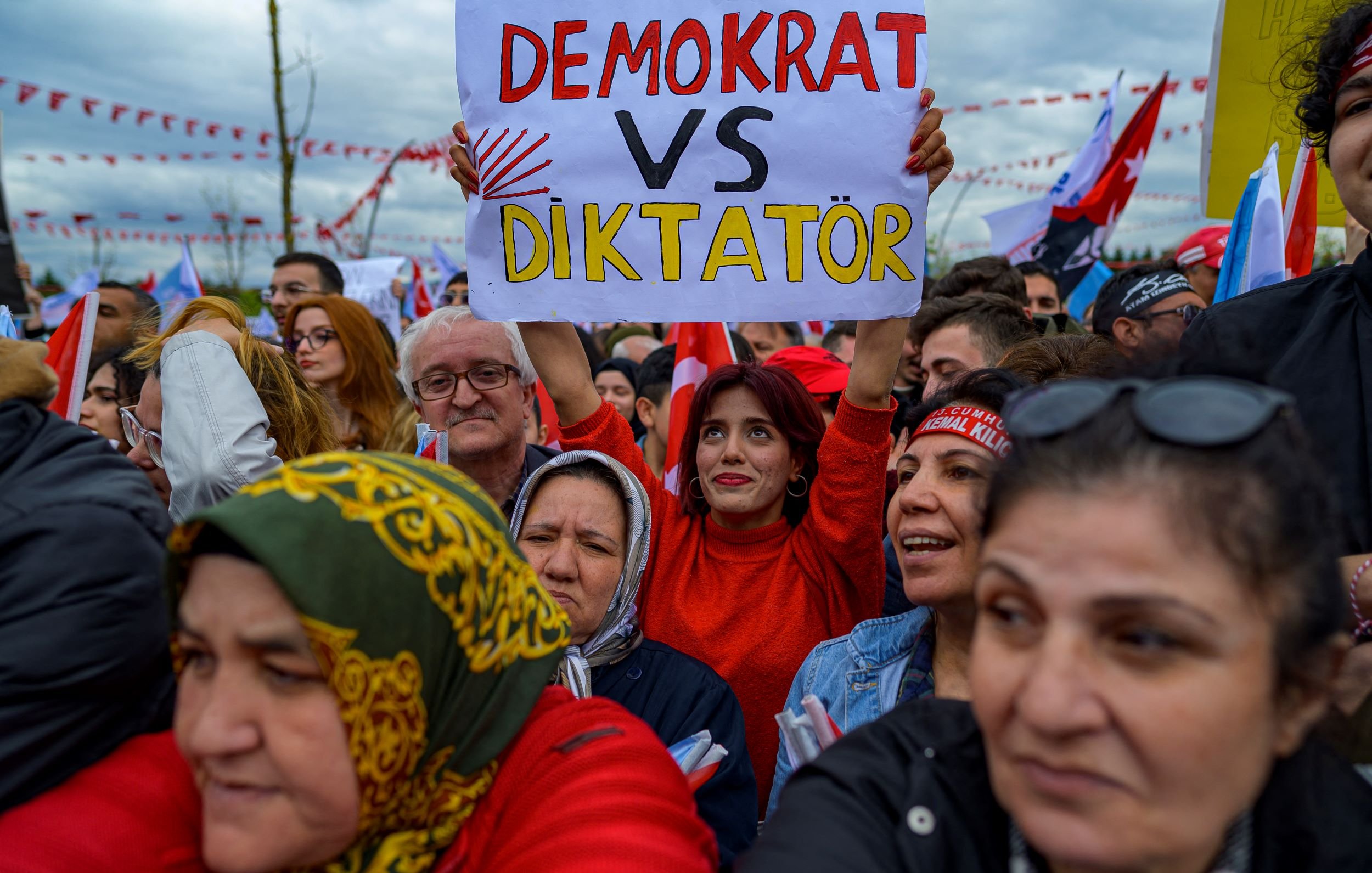 Nell'immagine si vede un gruppo di persone ad una manifestazione. In primo piano, una donna sorridente indossa una maglietta rossa e tiene in mano un cartello con la scritta "DEMOCRAT VS DIKTATÖR". Intorno a lei, ci sono diverse altre persone che sembrano partecipare attivamente all'evento. Molti di loro indossano sciarpe e cappelli colorati. Sullo sfondo, si vedono bandiere e decorazioni che contribuiscono all'atmosfera di protesta. L'ambientazione sembra carica di emozioni e impegno civico.