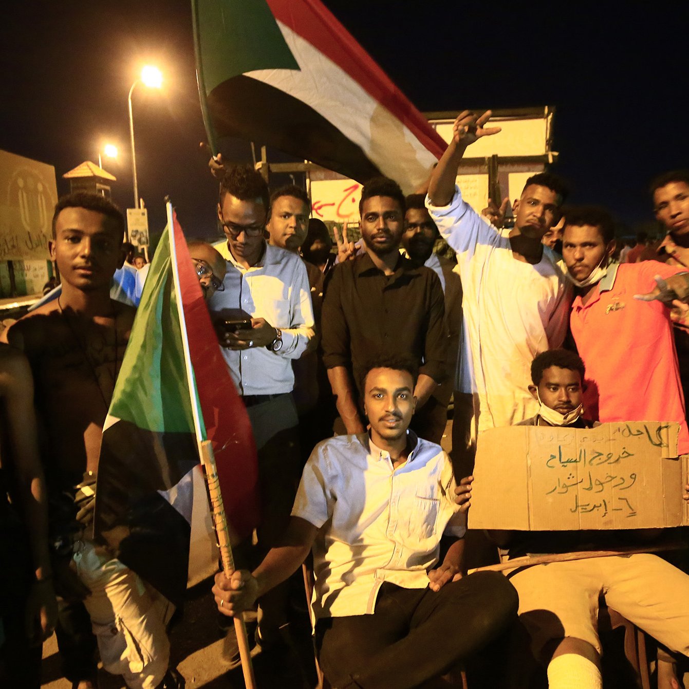 L'image montre un groupe de manifestants soudanais rassemblés lors d'une nuit, tenant des drapeaux et des pancartes. Certains d'entre eux sont debout, affichant des signes de solidarité avec les bras levés, tandis que d'autres sont assis, tenant des panneaux de revendication. L'atmosphère semble énergique et déterminée, incarnant un esprit de protestation et de demande de changement. Les visages des participants expriment diverses émotions, allant de la détermination à l'espoir.