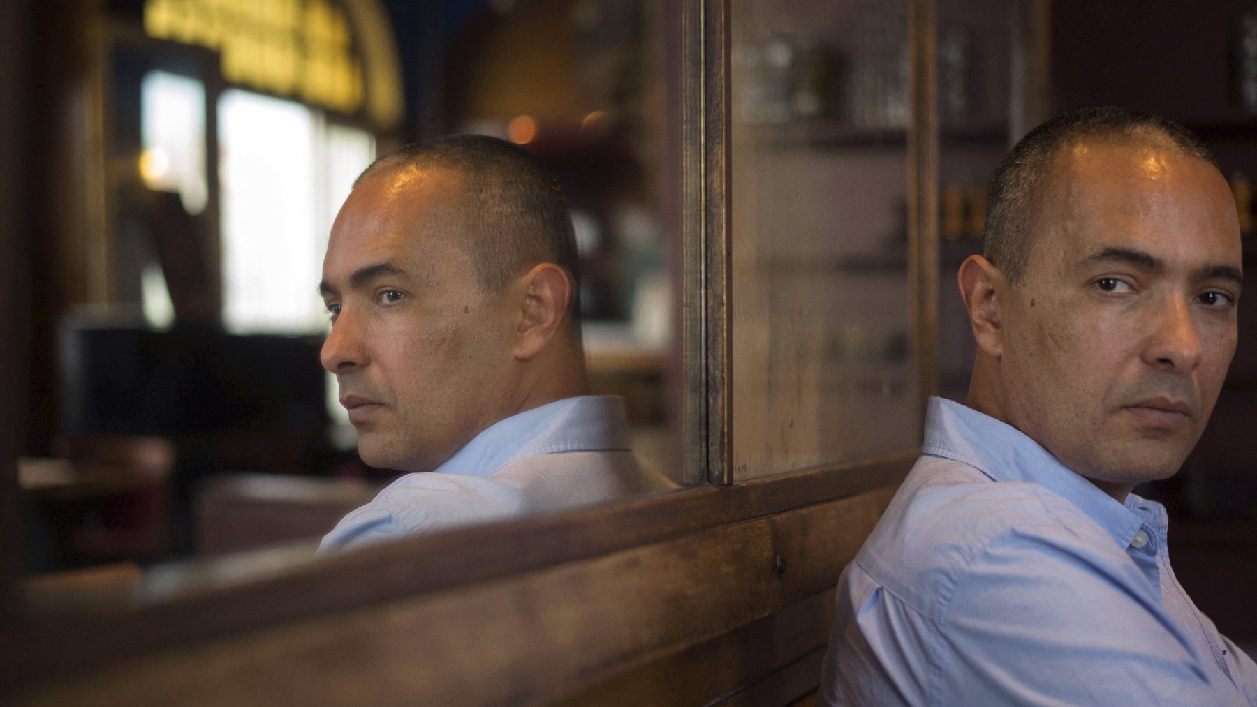 L'image montre un homme assis de dos, regardant vers un miroir qui reflète son visage. Il a une expression sérieuse, avec un léger froncement de sourcils. L'éclairage est doux et met en valeur les détails de son visage ainsi que l'ambiance de l'endroit où il se trouve, qui semble être un café ou un restaurant. Le décor en arrière-plan est flou, ce qui attire l'attention sur l'homme et son reflet.