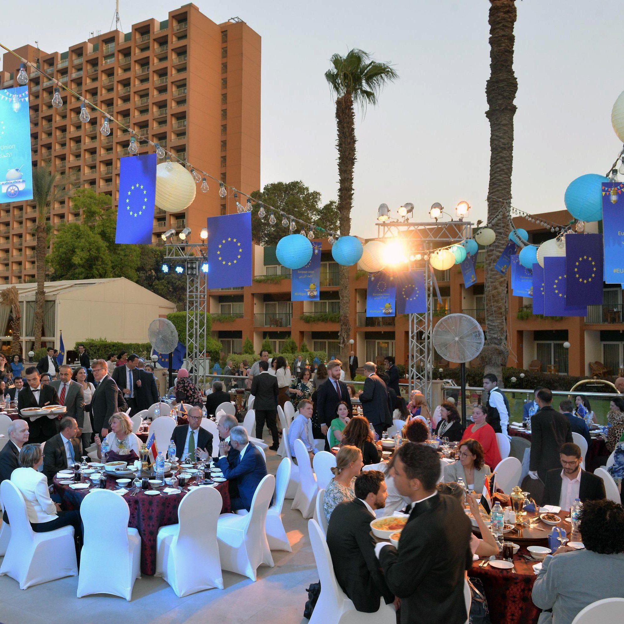 La imagen muestra un evento al aire libre, posiblemente una cena o celebración formal. Hay varias mesas decoradas con manteles elegantes y sillas blancas. Los asistentes, vestidos con ropa formal, están conversando y disfrutando de la comida. En el fondo, se pueden ver palmeras y un hotel o edificio alto. También hay decoraciones con banderas que parecen representar a la Unión Europea. Las luces brillantes y las lámparas de papel añaden un ambiente festivo a la escena. El clima parece agradable, sugiriendo que es una celebración durante la tarde o el atardecer.