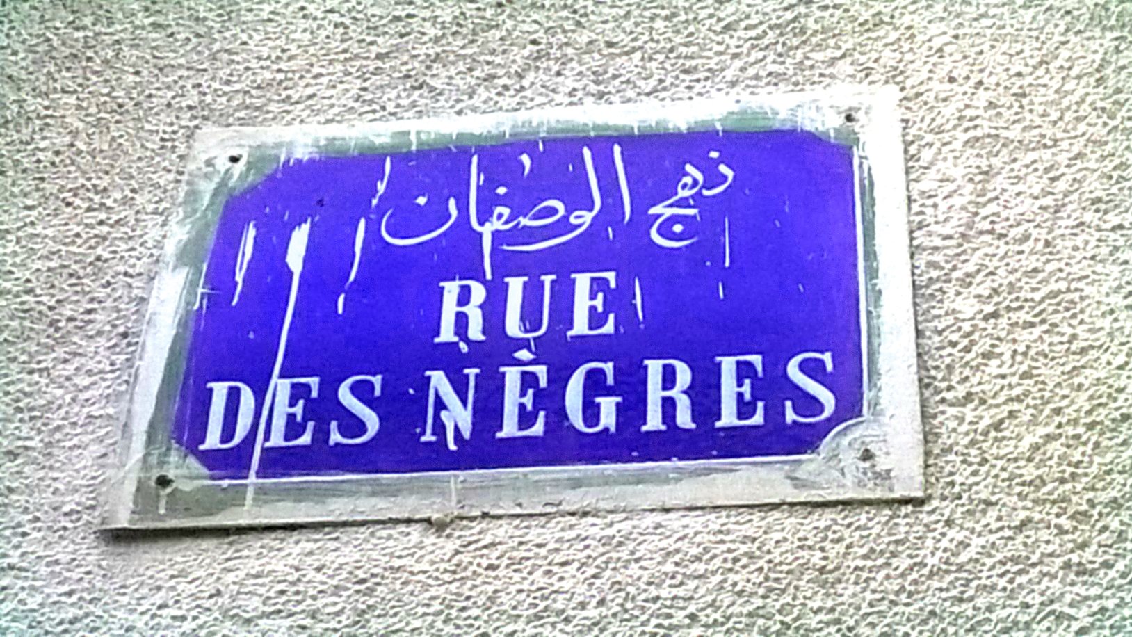L'image montre une plaque de rue. Elle est de forme rectangulaire et de couleur bleue. Le texte inscrit est en français et en arabe. En français, il est écrit "RUE DES NÈGRES", tandis qu'en arabe, le nom de la rue est également présent. La plaque semble un peu usée, avec des marques visibles comme de l'humidité ou des éclaboussures, ce qui indique qu'elle est probablement exposée aux éléments depuis un certain temps.