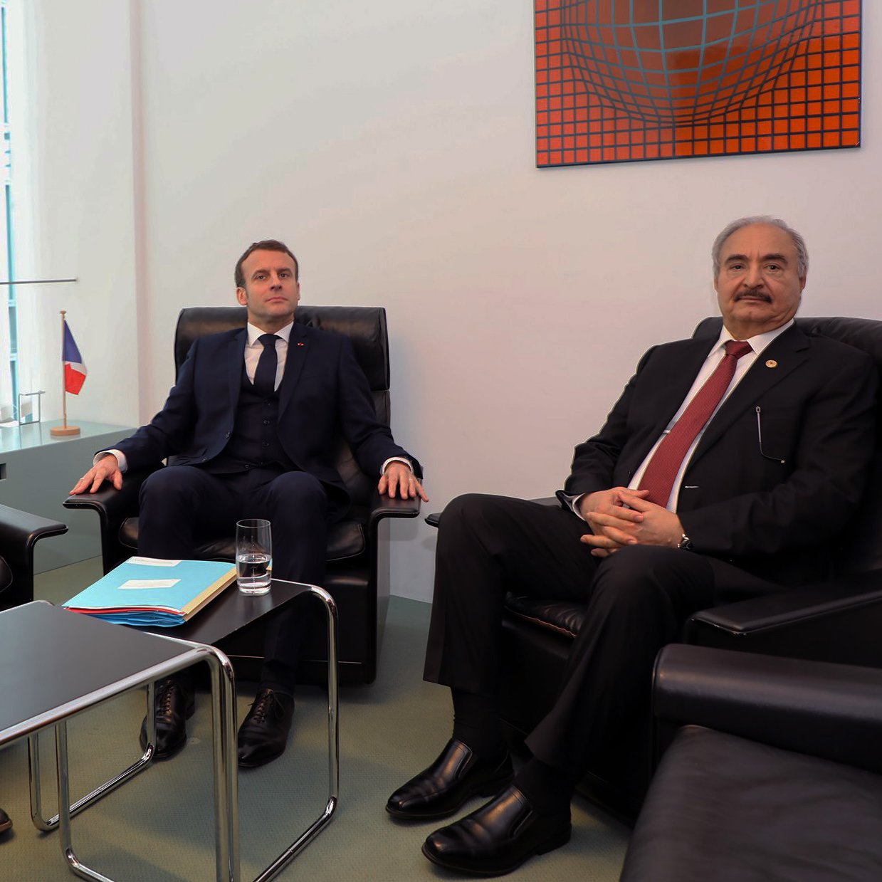 تظهر الصورة ثلاثة رجال يجلسون في غرفة اجتماعات. الشخص في المنتصف يرتدي بدلة داكنة، بينما الأخران يجلسان على جانبيه. يوجد علم فرنسي صغير في الجهة اليسرى من الصورة، والطاولة أمامهم تحتوي على أوراق وزجاج ماء. الجدران محاطة بالزجاج وتحتوي على عمل فني ملون في الأعلى. تعكس التعبيرات والوضوح في الجلسة وجود نقاش أو اجتماعات رسمية.