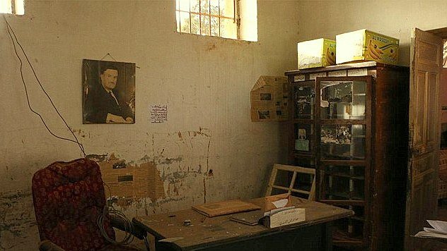 تظهر الصورة غرفة مهجورة مع جدران تحمل آثار الزمن، حيث تتواجد أريكة حمراء تقليدية على الجانبين. في الزاوية، يوجد مكتب خشبي بسيط بجانبه بعض الأوراق. على الجدار، توجد صورة معلقة لرجل يرتدي بدلة، مما يشير إلى أنه كان شخصية مهمة في الماضي. في الخلفية، يوجد زوج من خزائن الزجاج التي تحتوي على أشياء غير محددة. الإضاءة في الغرفة خافتة، مما يضيف إلى الأجواء الكئيبة للمكان.