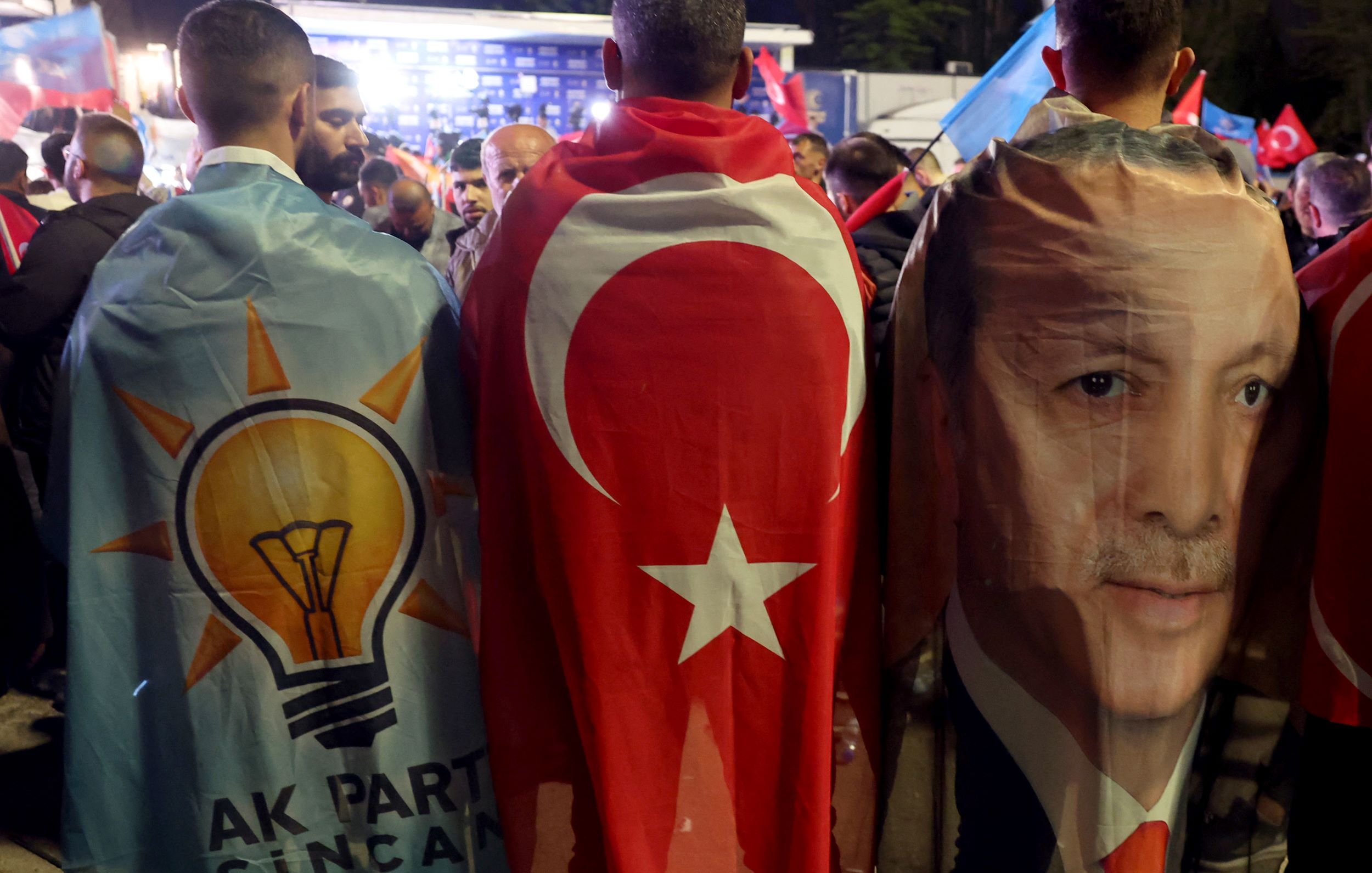 L'image montre un groupe de personnes réunies lors d'un événement politique, enveloppées dans des drapeaux et des bannières. Deux d'entre elles portent des capes : l'une présente le symbole du parti AKP (Parti de la justice et du développement), tandis que l'autre arbore le drapeau turc et une image du président Erdoğan. On peut voir une ambiance de rassemblement avec des drapeaux turcs en arrière-plan, suggérant une atmosphère de soutien et de célébration.