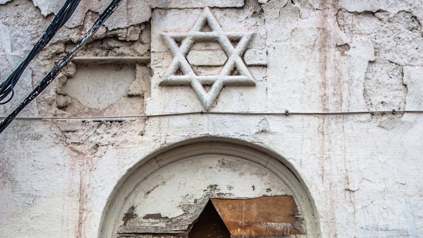 L'image montre un mur vieilli et abîmé, d'où une étoile de David est visible en haut, gravée ou ornée sur la surface. En dessous, une niche ou un cadre semble avoir été endommagé, avec des éléments en bois en mauvais état, suggérant un sentiment d'abandon. Un morceau de tissu bleu pend à l'intérieur de cette structure, ajoutant une touche de couleur à l'ensemble terni du mur. Les détails de la peinture écaillée et des fissures renforcent l'impression de délabrement et d'histoire.