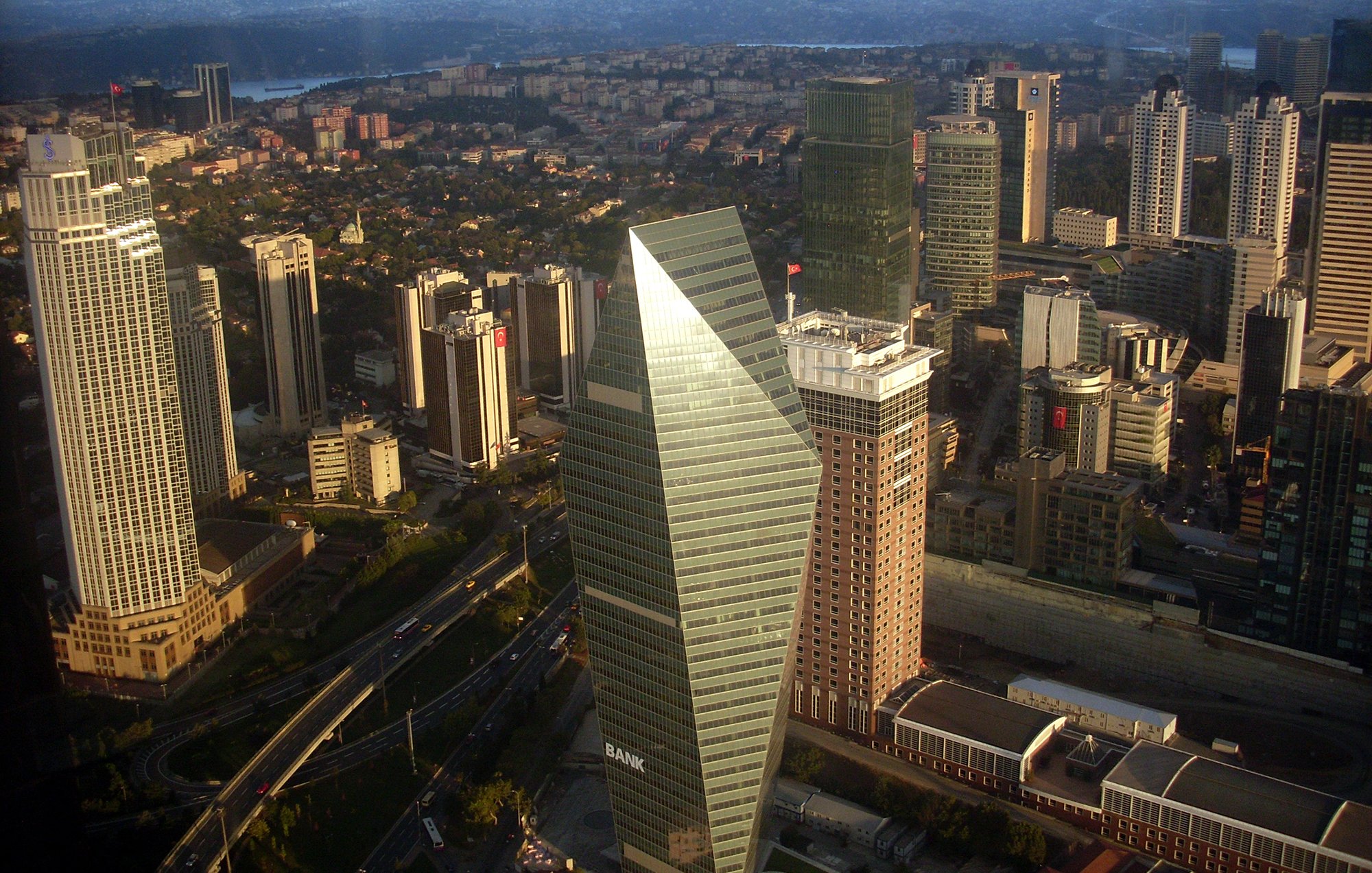 L'image montre une vue aérienne d'une métropole moderne, probablement une grande ville avec des gratte-ciels. On observe des bâtiments de différents styles architecturaux, certains avec des façades en verre réfléchissant. Au centre, un bâtiment en forme de pyramide avec des bords angulaires se détache, portant le mot "BANK". La ville est entourée de collines et d'un ciel partiellement nuageux, ce qui indique une atmosphère dynamique. Les routes serpentent à travers les bâtiments, suggérant une activité urbaine animée.