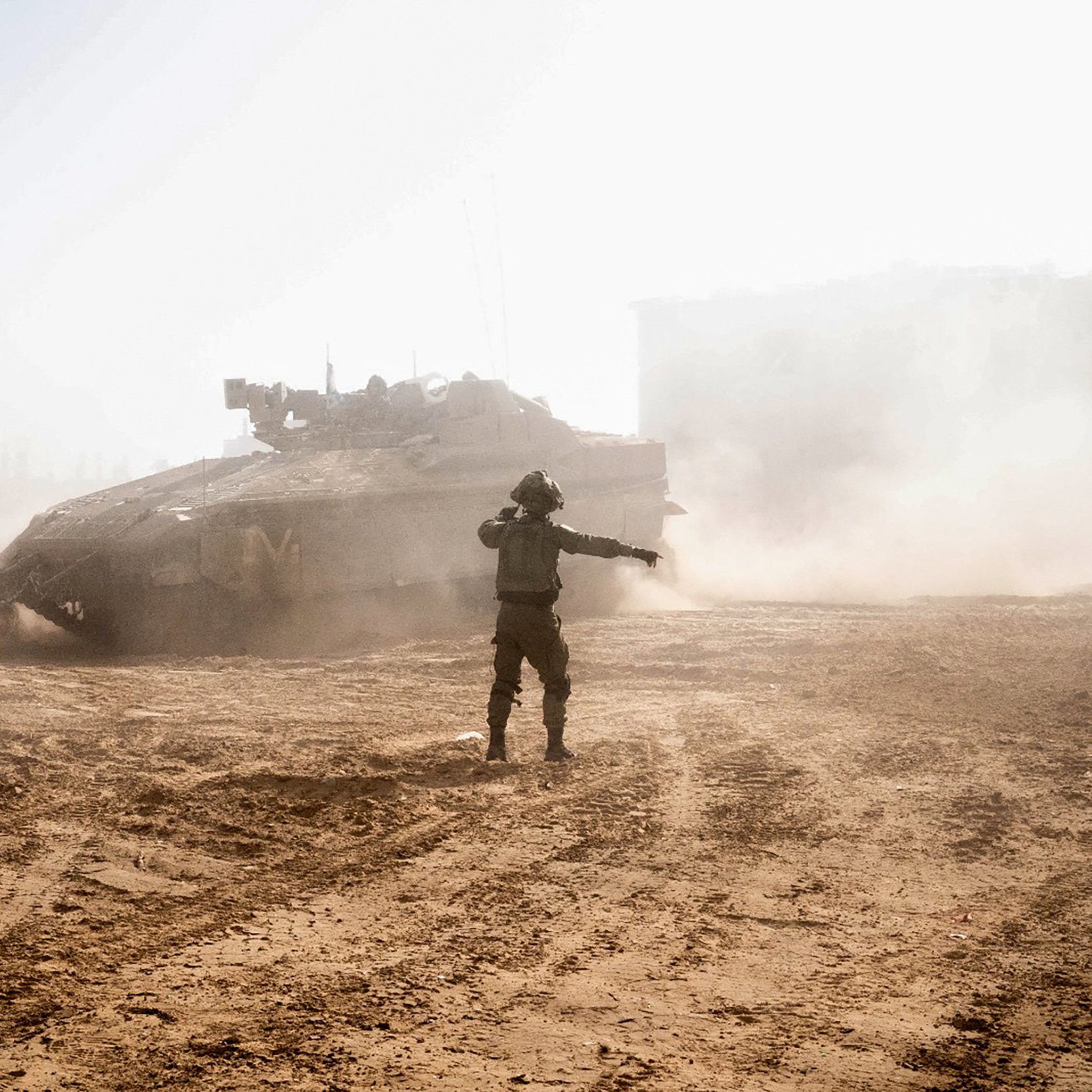 L'immagine mostra un soldato che si trova in un terreno polveroso, mentre un carro armato si muove sullo sfondo. Il soldato sembra indicare qualcosa, con una postura che suggerisce una direzione o un comando. La scena è caratterizzata da una luce intensa, che crea un'atmosfera di tensione, e ci sono nuvole di polvere sollevate dal carro armato, contribuendo a un senso di movimento e caos. Sullo sfondo, si intravede una struttura, ma i dettagli non sono ben definiti a causa della luce e della polvere.