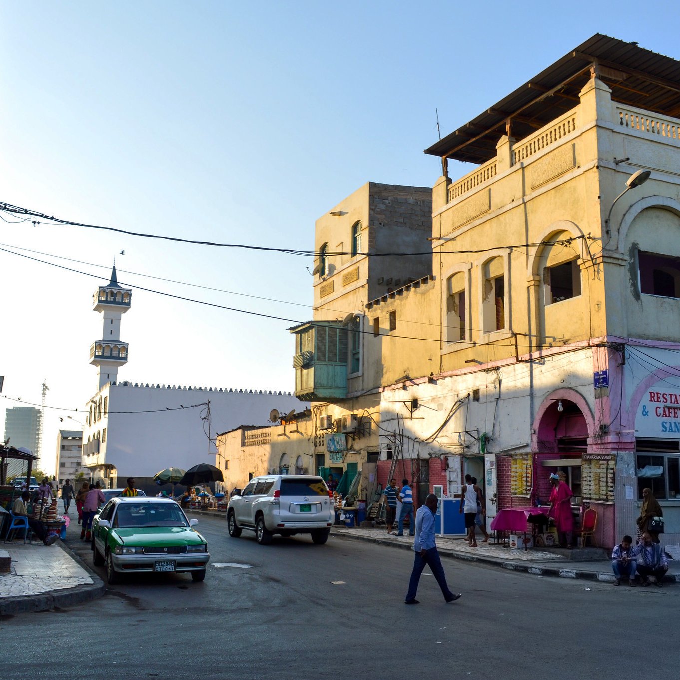 L'image montre une scène de rue animée dans une ville. On peut voir des bâtiments aux façades jaunes et blanches, typiques de l'architecture locale. À gauche, il y a des boutiques et un restaurant avec une enseigne bien visible. Sur la droite, un homme traverse la rue, entouré de véhicules stationnés et circulant. En arrière-plan, un minaret se dresse, indiquant la présence d'une mosquée. L'atmosphère est vivante, avec des passants et des couleurs vives qui caractérisent cet environnement urbain.