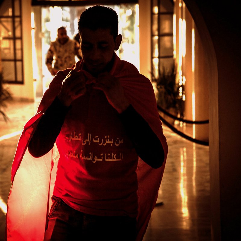 L'image montre un homme portant un t-shirt rouge avec un drapeau. Il semble s'apprêter à sortir, avec une ambiance chaleureuse et lumineuse en arrière-plan. On peut voir des éléments de décor qui suggèrent un espace intérieur, avec des silhouettes floues et une lumière dorée qui crée une atmosphère dramatique. L'homme porte une expression sérieuse et déterminée.