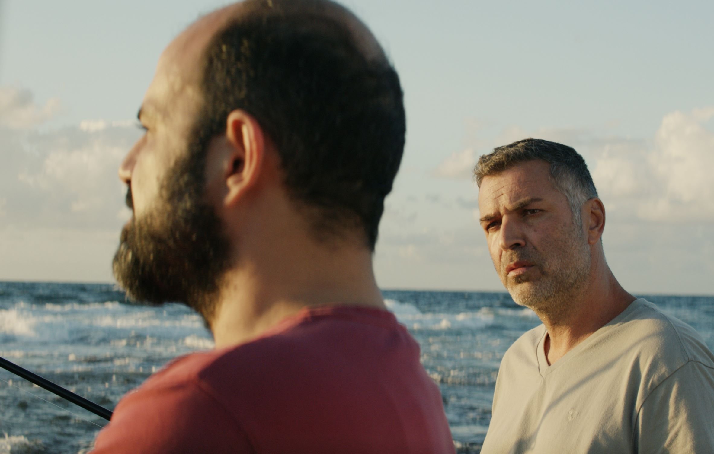 L'image montre deux hommes se tenant près de la mer. L'un d'eux a une barbe et porte un t-shirt rouge, tandis que l'autre a les cheveux grisonnants et un t-shirt clair. L'arrière-plan présente un océan calme sous un ciel bleu parsemé de nuages. L'atmosphère semble sereine, mais les expressions des hommes suggèrent une conversation intense ou réfléchie.