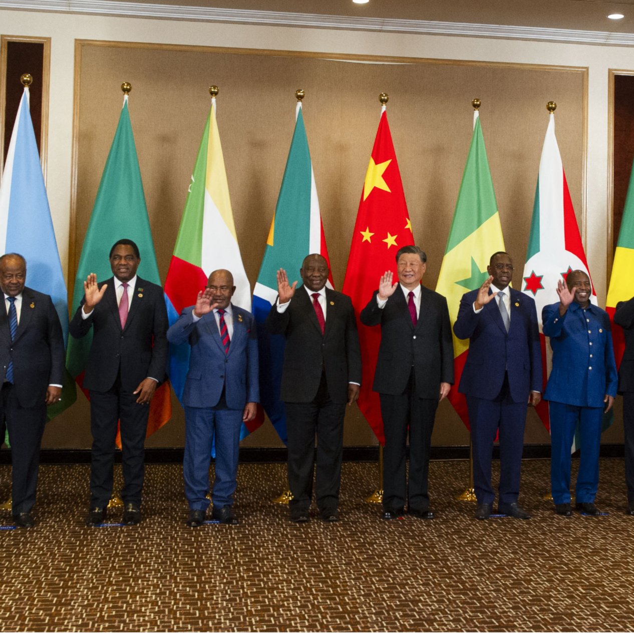 L'image montre un groupe de personnes debout en rangée, saluant. Elles se trouvent devant une série de drapeaux représentant différents pays. Les personnes portent des costumes formels et semblent participer à un événement diplomatique ou à un sommet international. Les drapeaux en arrière-plan incluent ceux de l'Union africaine, de la Chine, ainsi que d'autres nations. L'ambiance générale semble conviviale et respectueuse.