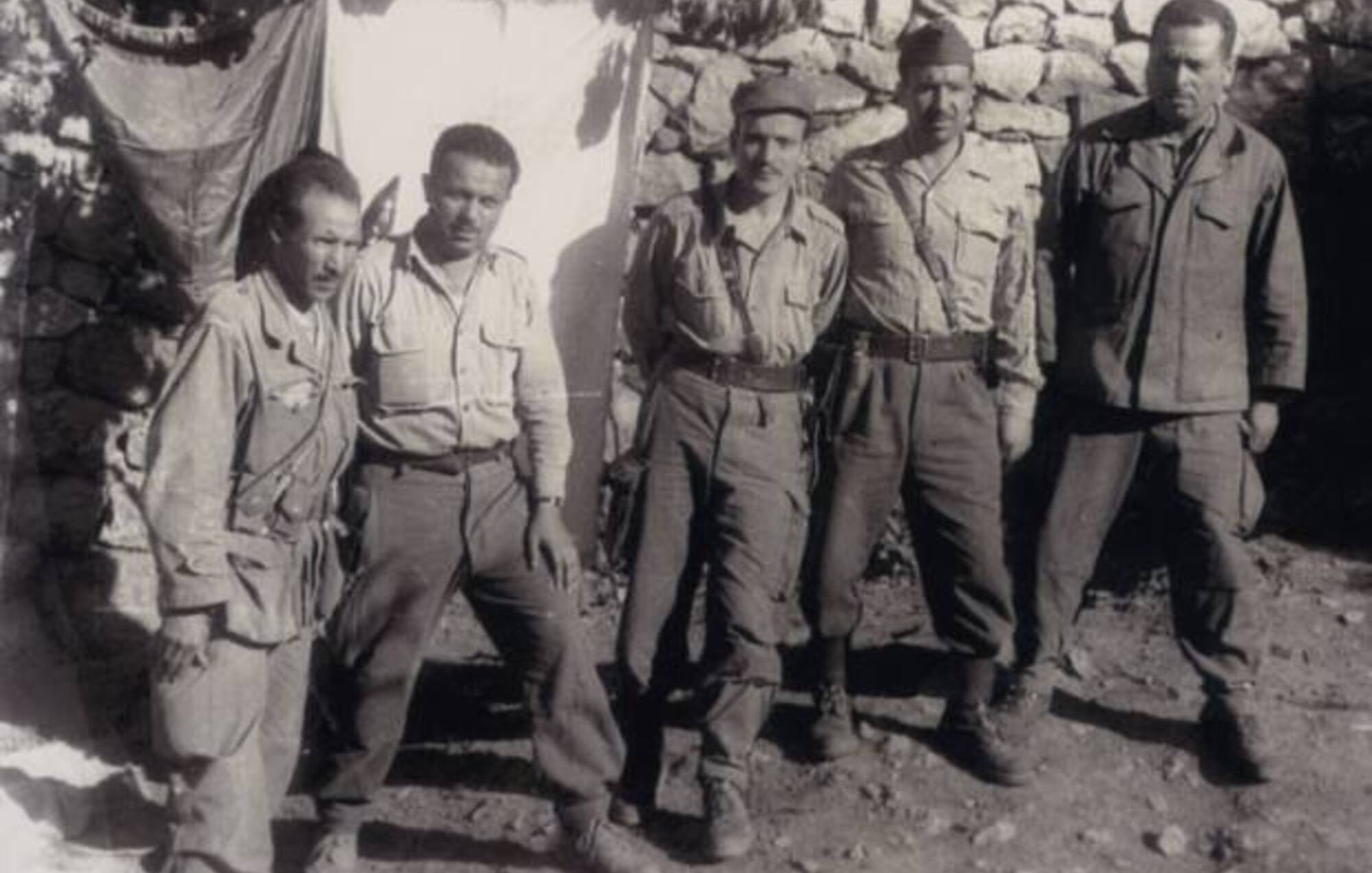 L'image présente un groupe de cinq hommes en uniforme militaire, debout ensemble devant un mur en pierres. Les hommes semblent être d'origine variée, portant des vêtements similaires, avec des pantalons et des chemises. Deux d'entre eux tiennent des armes à feu, et leur expression dégage un sentiment de camaraderie et de détermination. En arrière-plan, on aperçoit un drap suspendu, ce qui suggère qu'ils se trouvent dans un camp ou un endroit de rassemblement. L'atmosphère est sérieuse, typique des contextes militaires.