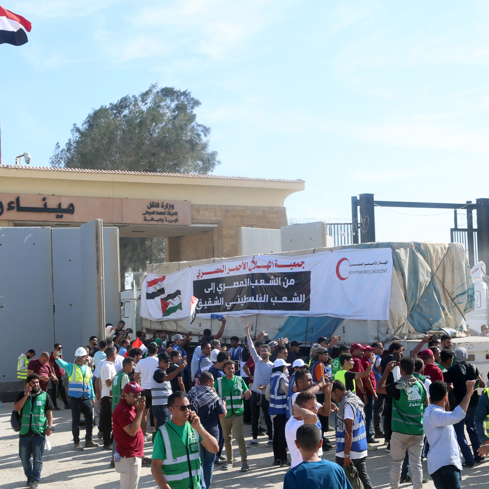 L'image montre une scène de manifestation à un point de passage, probablement en rapport avec des événements politiques. On peut voir des gens rassemblés, certains portant des gilets fluorescents, et une grande banderole avec un message en arabe. Des drapeaux, dont celui de l'Égypte, flottent à proximité. Le cadre semble être une zone de contrôle ou un poste de frontière, et l'ambiance est animée avec des personnes qui discutent ou manifestent.