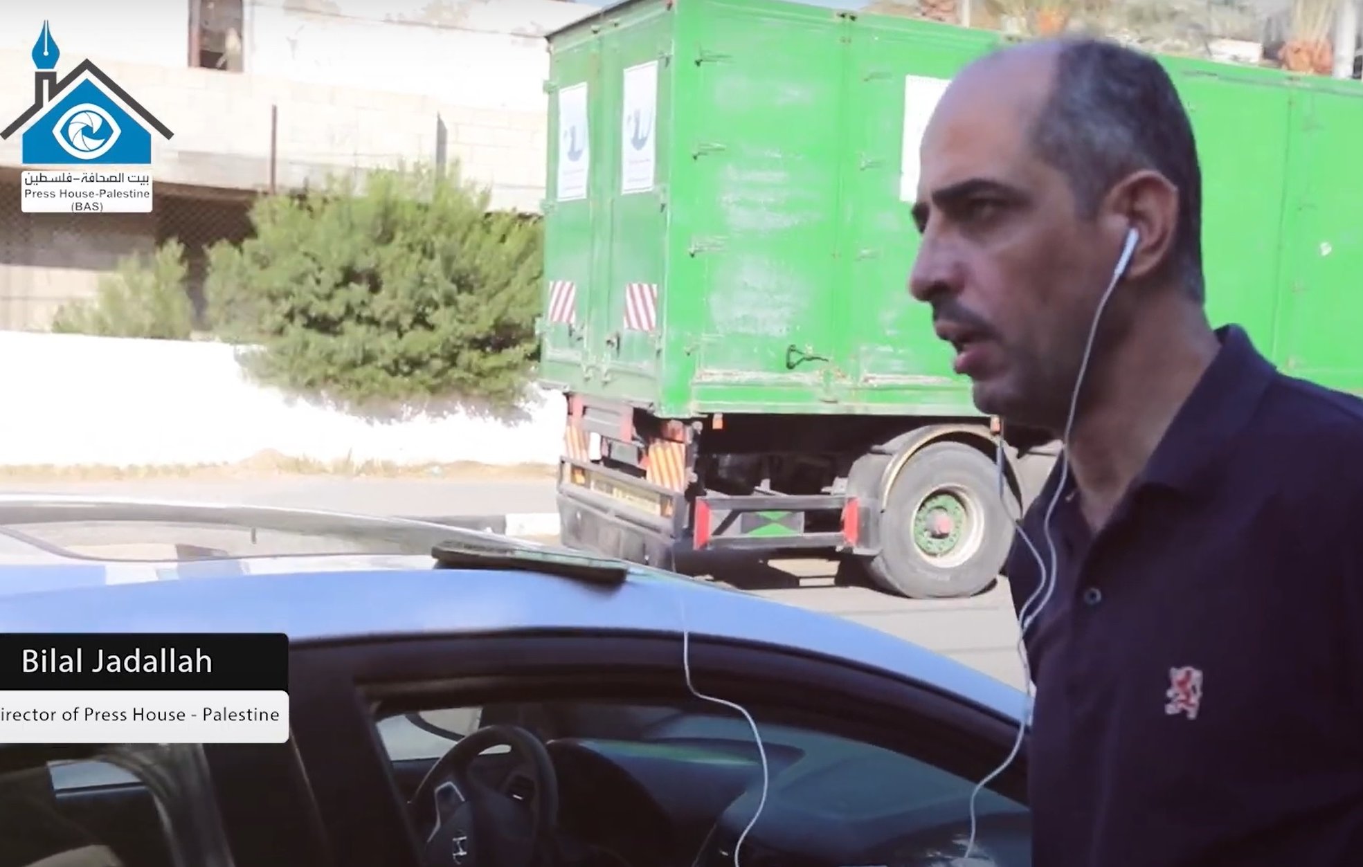 L'image présente un homme marchant à côté d'une voiture, portant des écouteurs. Il a un air concentré et semble être dans un environnement urbain avec un camion vert en arrière-plan. Un texte en bas de l'image mentionne "Bilal Jadallah, Directeur de Press House - Palestine".