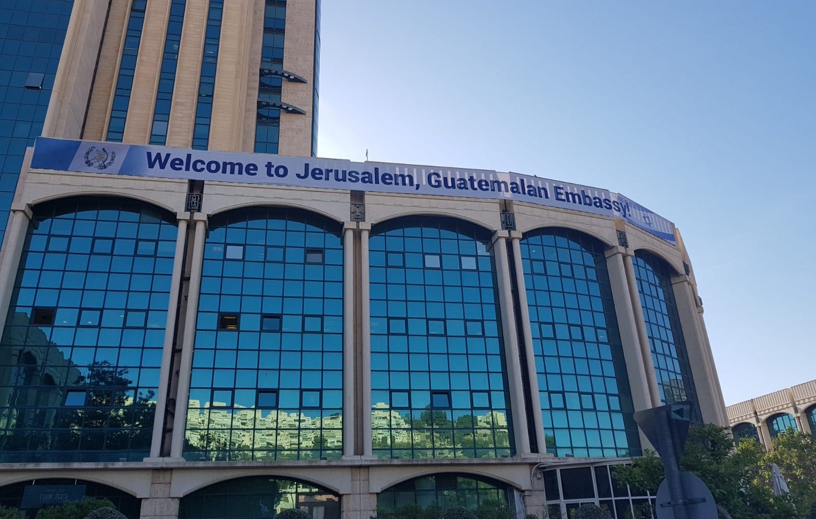L'immagine mostra un edificio con una grande facciata di vetro riflettente. Sulla parte superiore dell'edificio è affisso uno striscione che dice "Welcome to Jerusalem, Guatemalan Embassy". Il contesto suggerisce che si tratta della sede dell'ambasciata del Guatemala a Gerusalemme. Si può notare il cielo blu sullo sfondo e alcune piante o alberi nella parte inferiore dell'immagine.