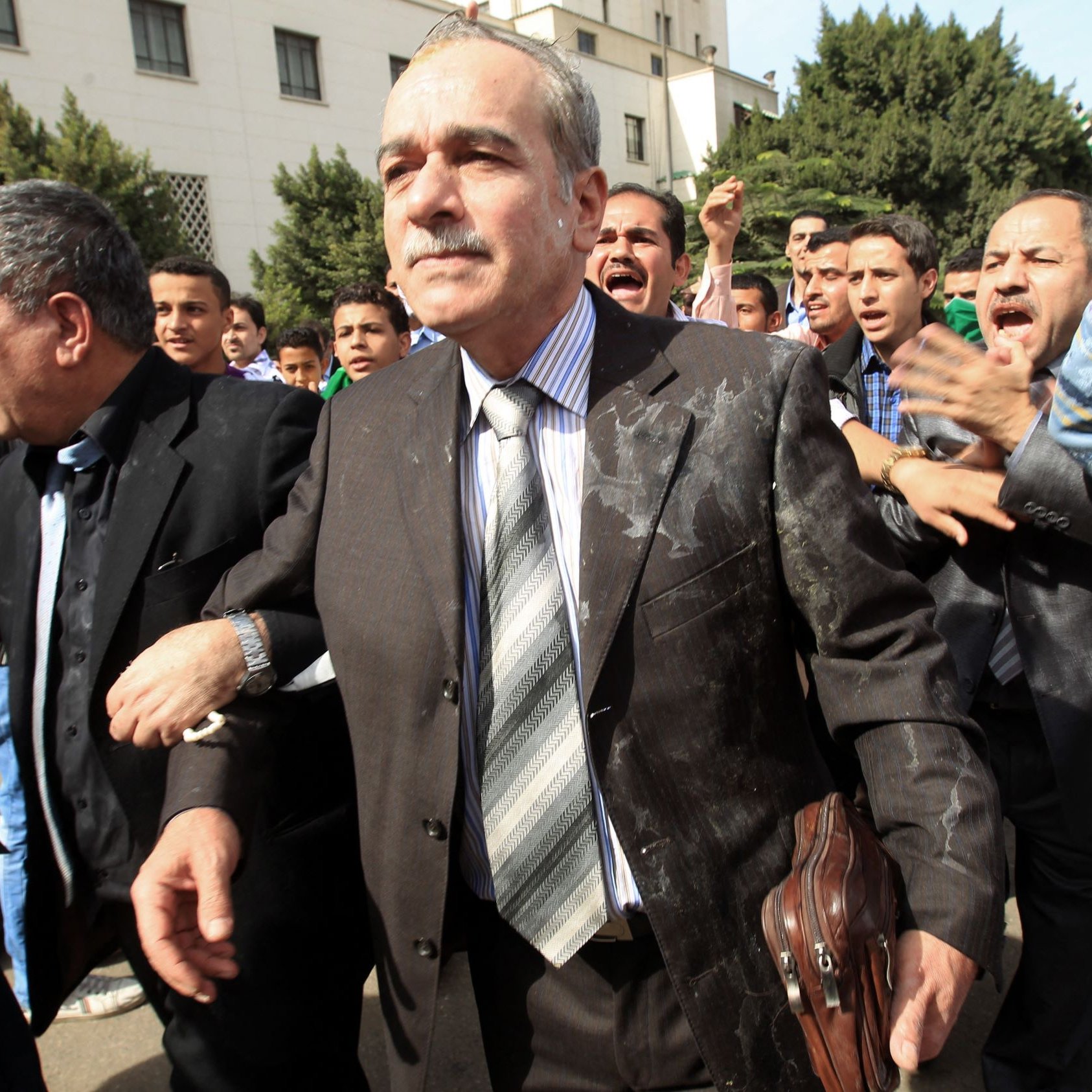 تظهر الصورة مجموعة من الأشخاص في مكان عام، حيث يبدو أن هناك تفاعل قوي ومثير. في المقدمة، يوجد رجل يرتدي بدلة رسمية ويظهر على وجهه تعبير جدّي. يحيط به عدد من الأشخاص الآخرين الذين يبدو أنهم متحمسون أو منفعلون، مما يوحي بوجود حدث مهم أو احتجاج. الخلفية تضم مجموعة من الأشجار والمباني، مما يعطي انطباعًا بأن المكان هو منطقة حضارية. الجو العام في الصورة يبدو ديناميكيًا ومليئًا بالطاقة.