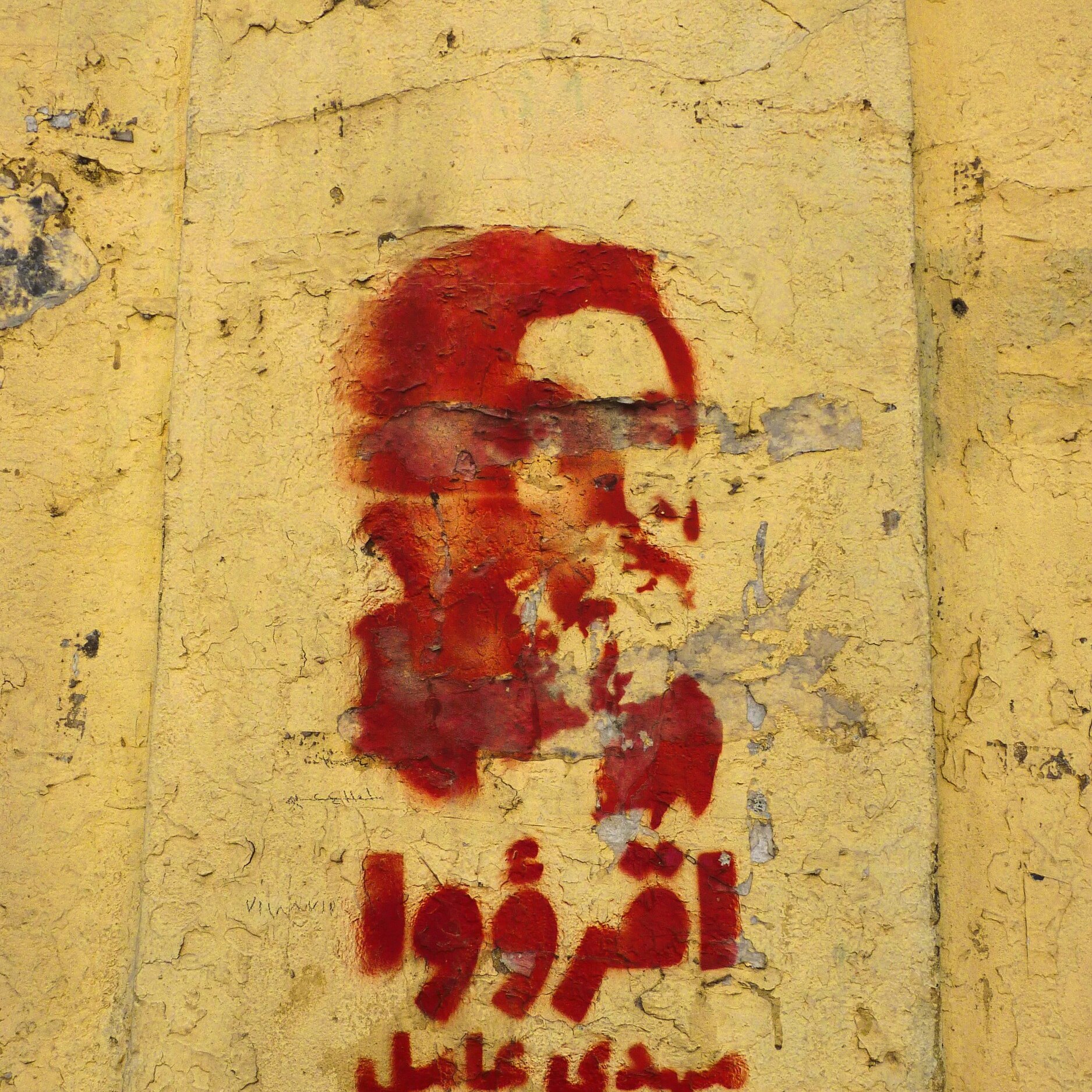 L'immagine mostra un murale su una parete di colore giallo. Il murale rappresenta un volto stilizzato, probabilmente un personaggio noto, dipinto in rosso. Sotto il volto ci sono delle scritte, anch'esse in rosso, che sembrano in arabo. La superficie della parete appare consumata e scrostata, contribuendo a un aspetto storico e di protesta.