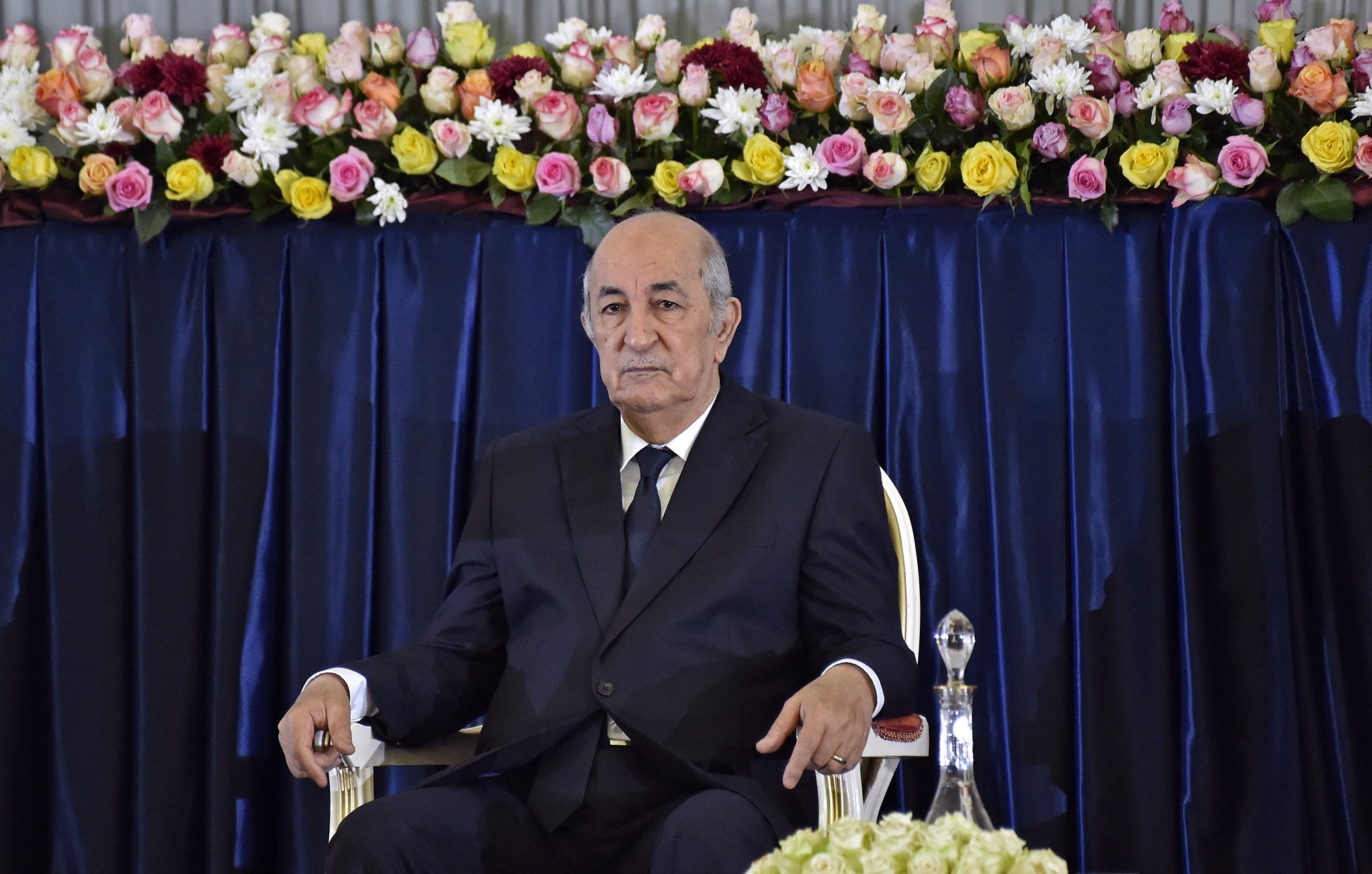 L'image montre un homme assis sur une chaise, portant un costume sombre et une cravate. Il a l'air sérieux et attentif. À l'arrière-plan, il y a une décoration florale composée de roses et d'autres fleurs qui ajoutent de la couleur à la scène. La disposition des fleurs est soignée, suggérant un cadre formel ou officiel.