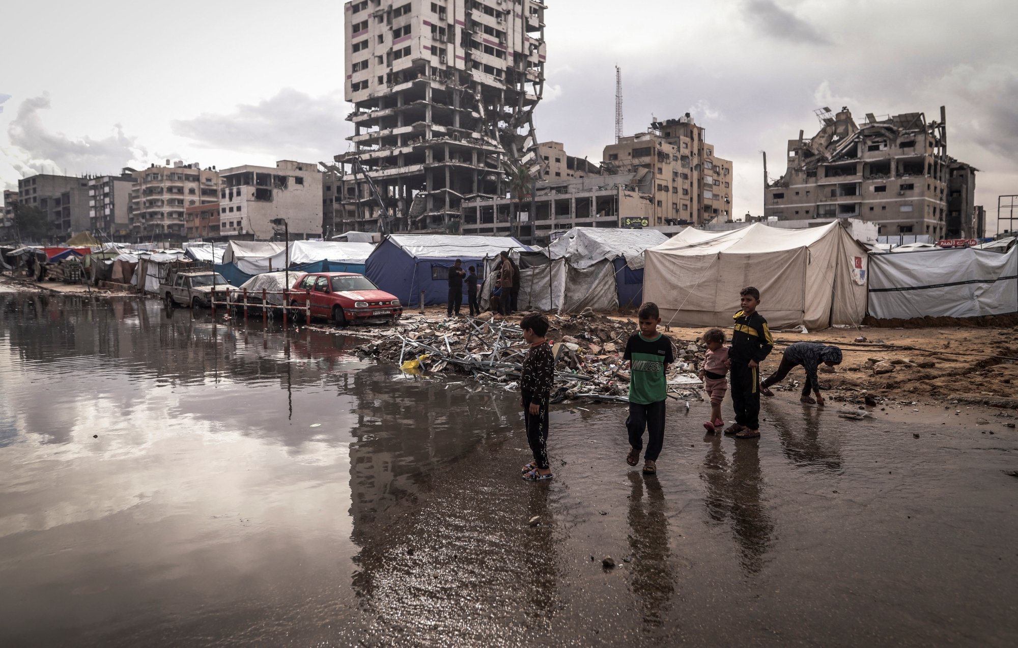 Des enfants jouent près de tentes dans un camp, avec un bâtiment en ruines en arrière-plan.