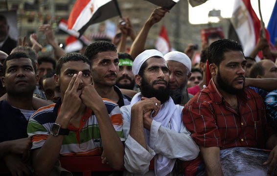 L'image montre un groupe de personnes rassemblées lors d'une manifestation. On peut voir des hommes de différentes origines, certains portant des vêtements traditionnels, d'autres des t-shirts colorés. Ils semblent tous attentifs et engagés, certains levant les mains ou tenant des drapeaux. L'ambiance semble être celle d'une mobilisation ou d'une expression de soutien pour une cause. Le fond montre un environnement urbain, avec d'autres manifestants et des drapeaux visibles.
