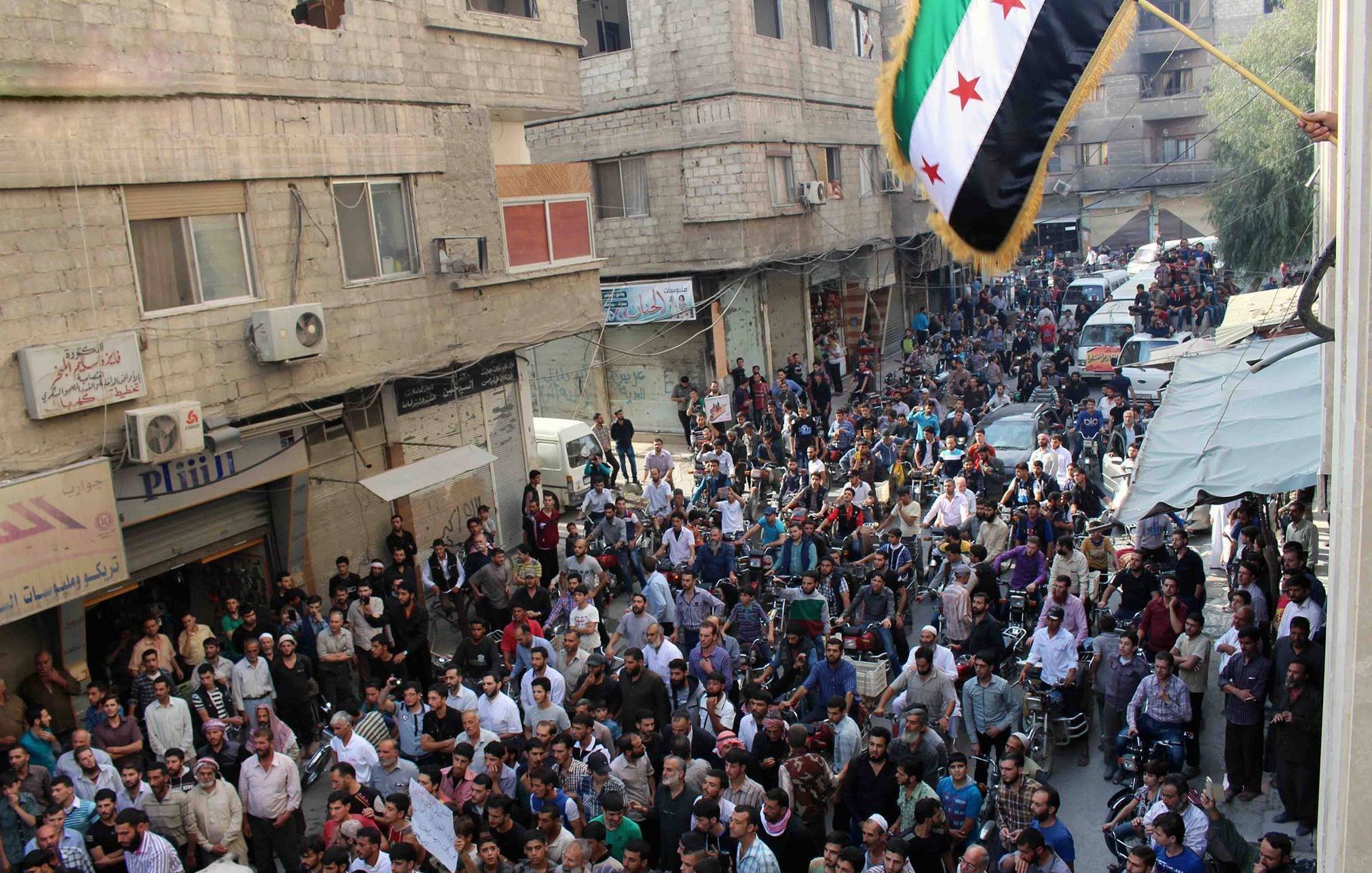 الصورة تظهر حشودًا كبيرة من الناس في شارع مزدحم. يتجمع المحتجون في منطقة حضرية، حيث يمكن رؤية المباني والأسطح والشرفات. علم يرفرف في الأعلى، وهو العلم السوري المعتمد من قبل بعض فصائل المعارضة. يبدو أن هناك جو من الحماس والتجمع، مع وجود بعض السيارات والمركبات في الخلفية.