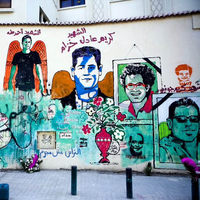 L'image montre un mur recouvert de fresques murales colorées. On y voit plusieurs portraits et illustrations. L'une des figures centrales semble représenter une personne avec des ailes, symbole souvent associé à un message d'espoir ou de mémoire. Des textes en arabe et en français peuvent être aperçus, évoquant des thèmes de la liberté et de la résistance. En bas, une personne se tient debout, probablement en train de peindre ou d'admirer l'œuvre. L'atmosphère générale dégage une impression vibrante et engagée, reflétant une culture urbaine riche.