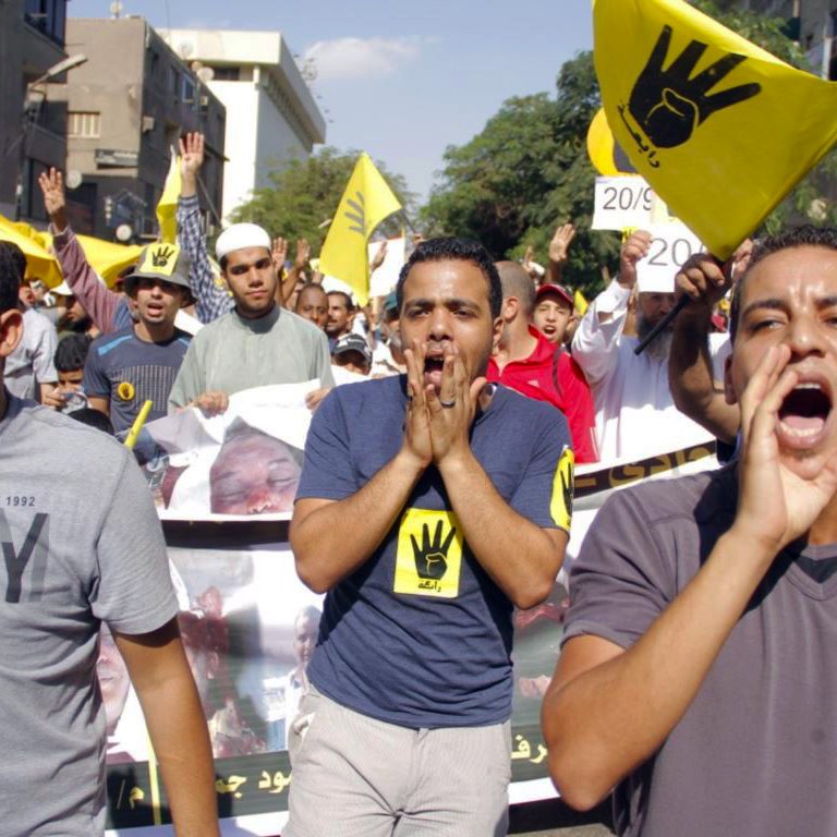 La imagen muestra a un grupo de personas participando en una manifestación. Los manifestantes parecen estar expresando su descontento, gritando y levantando las manos. En el fondo, se pueden ver pancartas y banderas, algunas de las cuales tienen un símbolo de mano levantada. El ambiente es de protesta, y la multitud está activa y unida en su mensaje. La escena se desarrolla en un entorno urbano, con edificios visibles en el fondo.