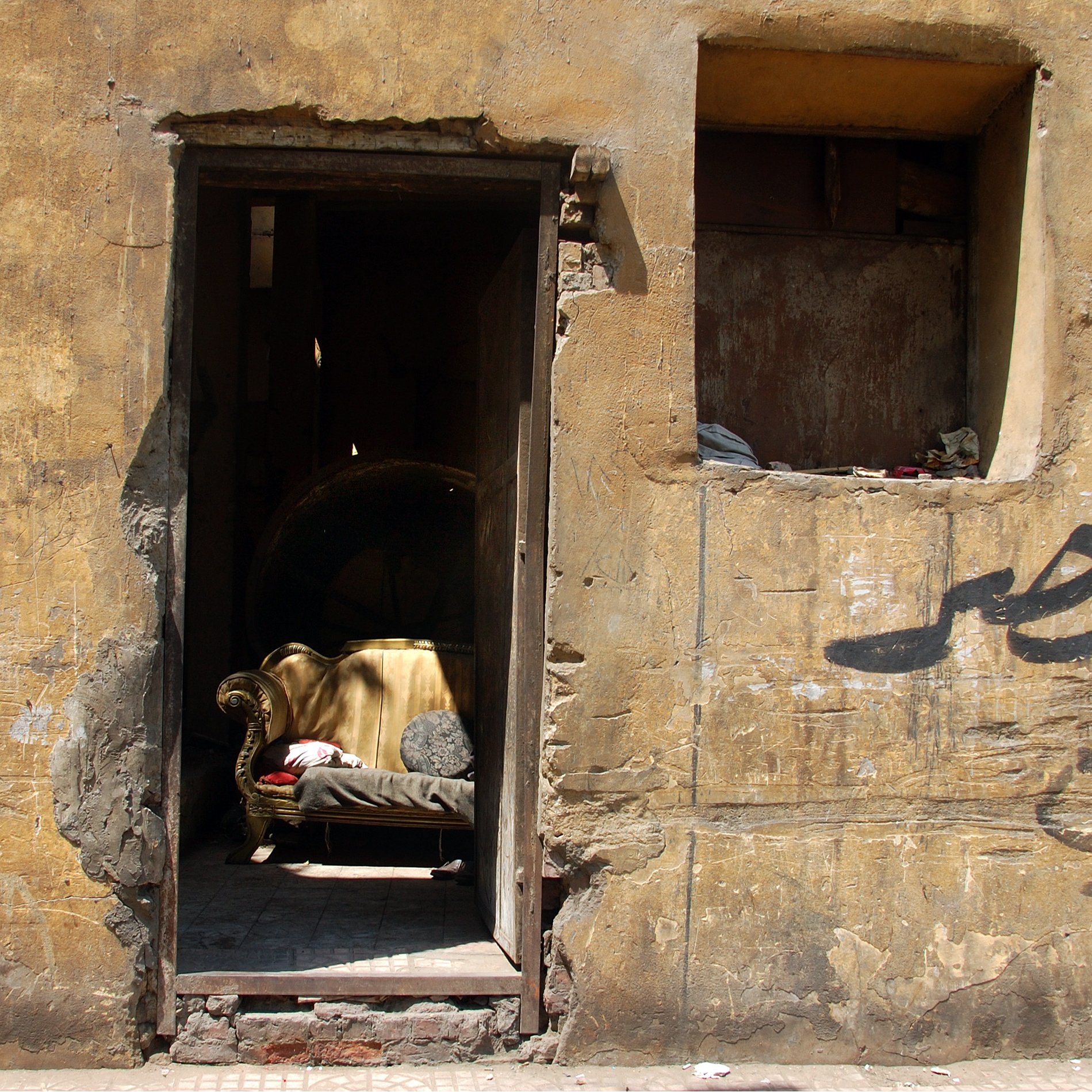 تصویر نمایی از یک درب ورودی به یک ساختمان قدیمی است که دیوارهای آن رنگ و بافت زرد و کهنه‌ای دارند. در وسط در، یک کاناپه قدیمی و ساده قرار دارد که به نظر می‌رسد سال‌ها بدون استفاده مانده است. در سمت چپ تصویر، بخشی از دیوار به صورت تخریب شده دیده می‌شود و یک پنجره کوچک بالای درب نیز وجود دارد که نیمی از آن به طور کامل تخریب شده است. روی دیوار نوشته‌هایی به زبان عربی دیده می‌شود که پس‌زمینه‌ای از فضای شهری و قدیم را به تصویر می‌کشد. کل صحنه حس نوستالژیک و متروکه‌ای را منتقل می‌کند.