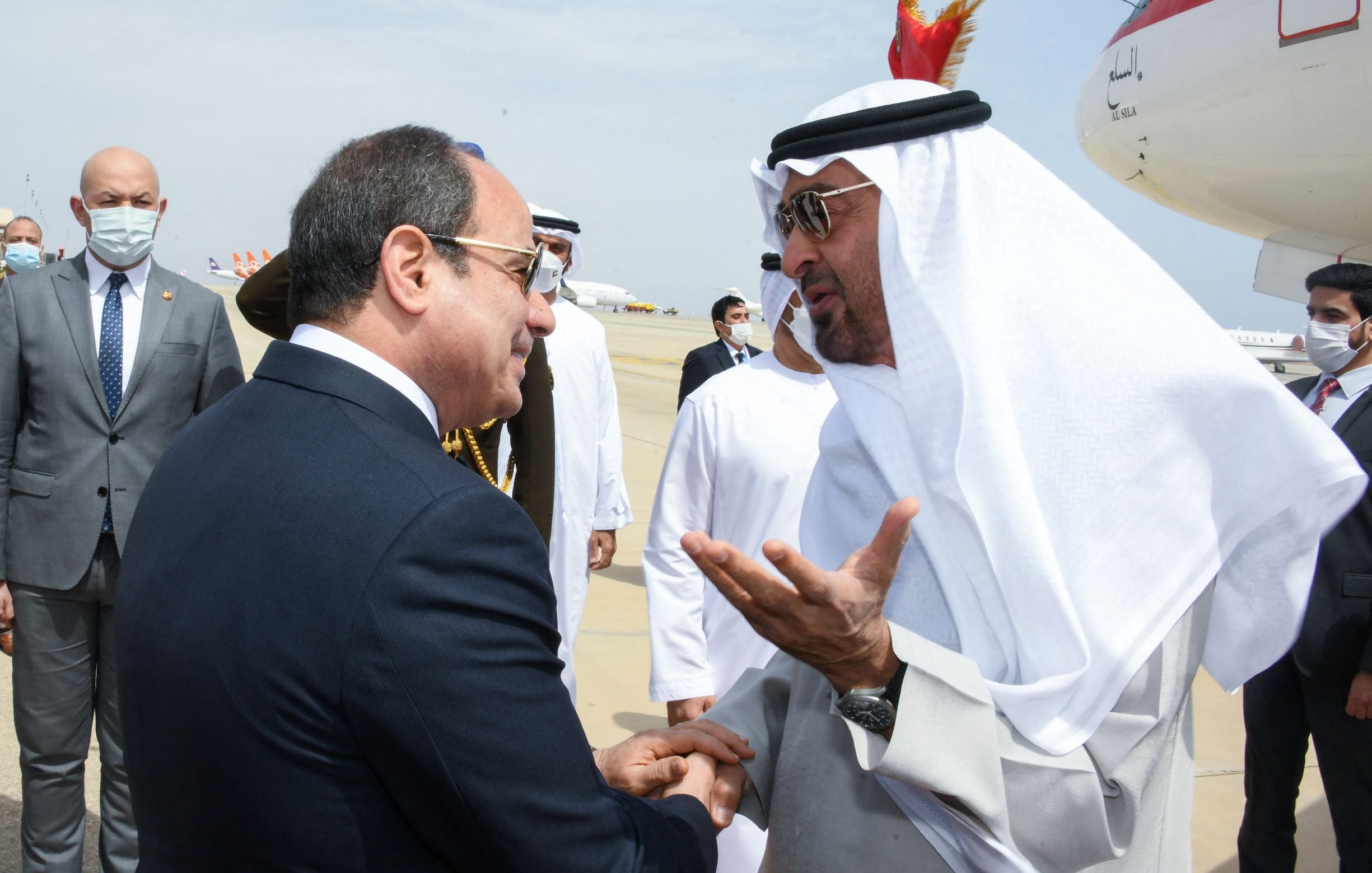 L'image montre une rencontre entre deux dirigeants. Ils semblent être en conversation amicale, avec des gestes visibles pour accompagner leurs paroles. L'un des deux est vêtu d'un costume formel, tandis que l'autre porte des vêtements traditionnels arabes, incluant un keffieh. En arrière-plan, on peut apercevoir des membres du personnel et des avions, suggérant que la scène se déroule dans un aéroport. L'atmosphère paraît conviviale et respectueuse.