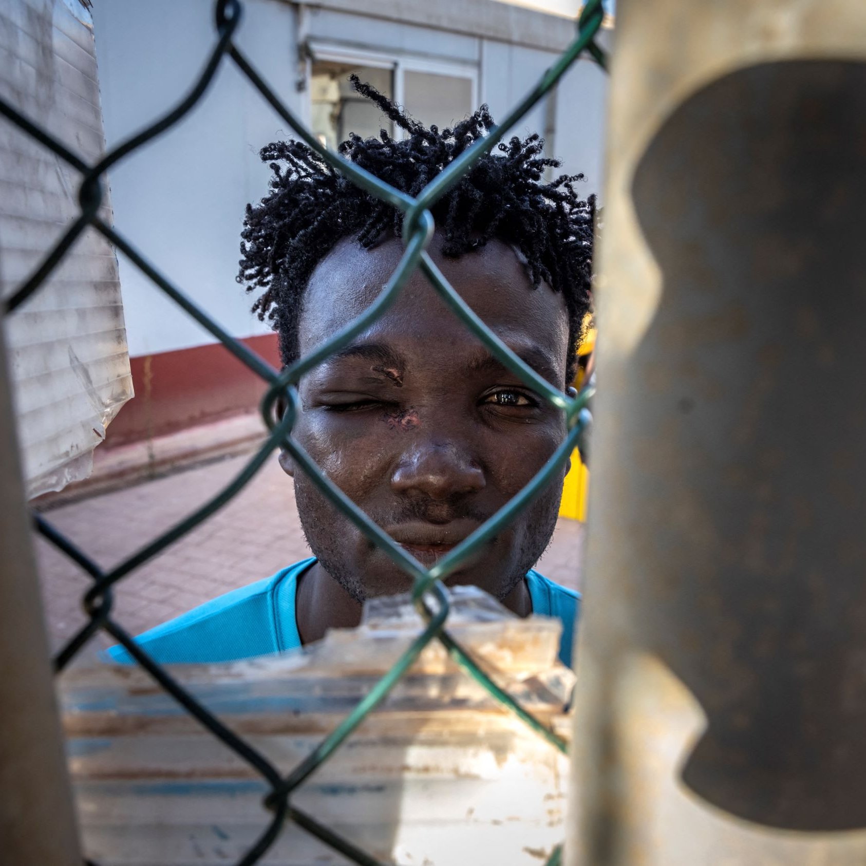 L'immagine mostra un giovane uomo che si affaccia attraverso una rete di recinzione. Ha i capelli ricci e un'espressione seria. Il suo viso è parzialmente coperto da una rete metallica, il che crea un effetto di separazione. Sullo sfondo, si intravede un'architettura semplice. La luce colpisce il suo volto, mettendo in evidenza i dettagli della sua espressione.