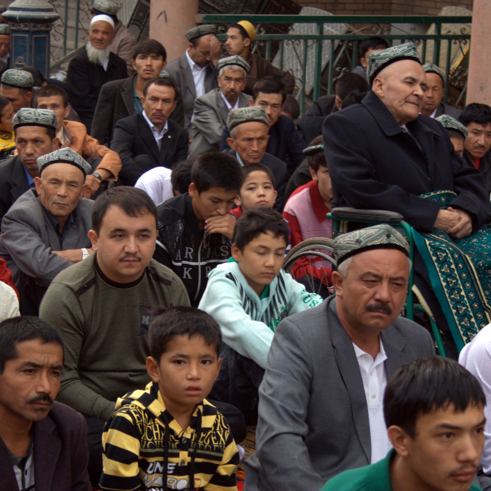 تظهر الصورة مجموعة من الأشخاص الجالسين في مكان مفتوح، يبدو أنهم مجتمعون في مناسبة أو تجمع. يوجد رجل مسن يجلس على كرسي متحرك في الخلف، ويبدو أنه يراقب ما يحدث من حوله. هناك تنوع في الأعمار بين الحضور، حيث توجد وجوه مراهقين وشباب وكبار في السن. يرتدي الكثير من الرجال قمصاناً تقليدية، وبعضهم يرتدي قُبُعات ذات تصاميم مميزة. يبدو أن الجو يسوده الانتباه والتركيز على الحدث الجاري.