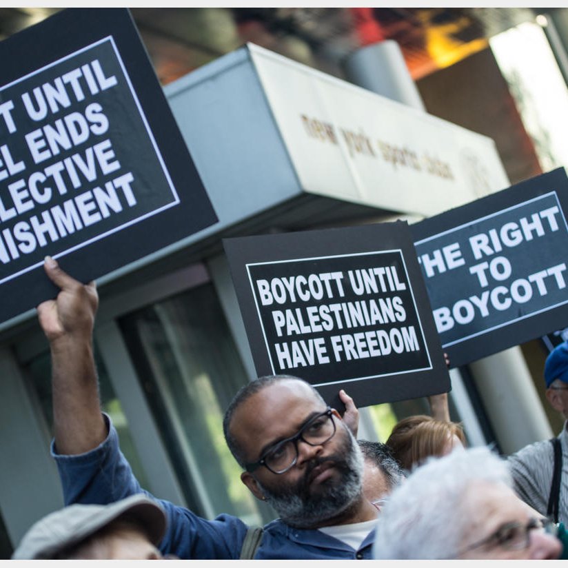 L'image montre une manifestation avec des personnes tenant des pancartes. Les pancartes portent des messages appelant au boycott jusqu'à ce qu'Israël mette fin à des mesures de punition collective et jusqu'à ce que les Palestiniens aient la liberté. On peut voir des participants de différents âges, tous unis par des revendications similaires. L'arrière-plan indique un environnement urbain avec des bâtiments.