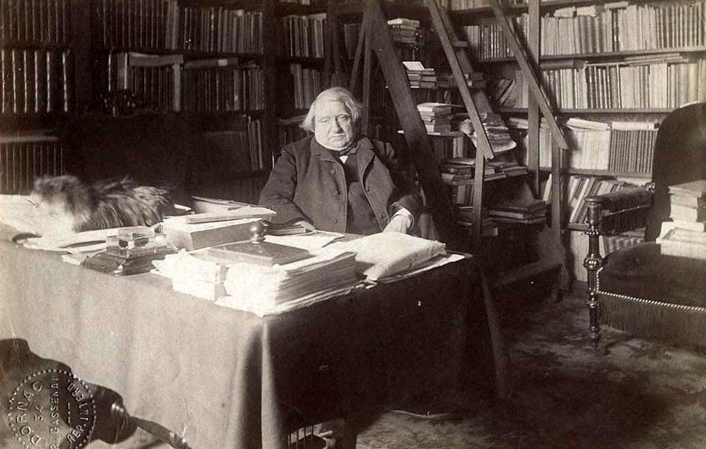 L'image montre une scène d'intérieur, probablement dans un bureau ou une bibliothèque. Un homme, apparemment âgé, est assis à une table chargée de papiers et de livres. Derrière lui, on peut voir des étagères remplies de livres. Il y a également un tapis sur le sol. Un chat, qui semble être sur la table ou à proximité, ajoute une touche de vie à l'atmosphère studieuse de la pièce. L'éclairage donne une impression de sérénité et de calme.