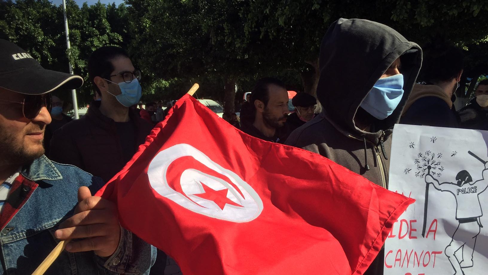 L'image montre un groupe de personnes participant à une manifestation. Au premier plan, un homme tient un drapeau tunisien rouge avec un croissant et une étoile blanche. D'autres manifestants portent des masques, et l'un d'eux tient une pancarte sur laquelle est écrit un message en anglais, notamment « IDEAS CANNOT BE DESTROYED ». L'arrière-plan est constitué d'arbres et de rues, ce qui donne un air de protestation pacifique.
