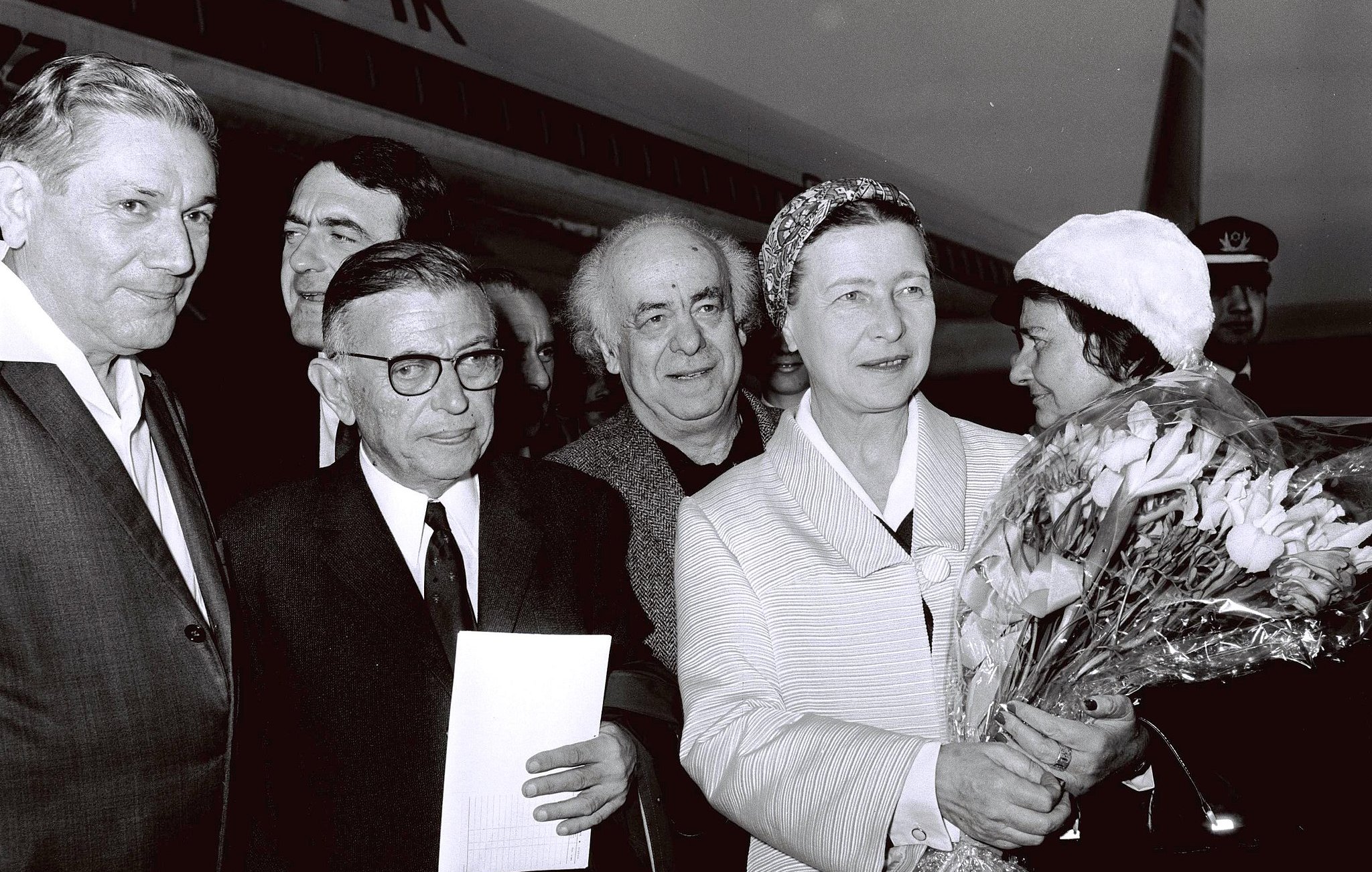 L'image montre un groupe de personnes qui accueillent une femme portant un bouquet de fleurs. Elle est entourée de plusieurs hommes, dont certains portent des lunettes, et tous semblent s'engager dans une conversation ou une interaction. L'environnement indique qu'ils sont à l'aéroport, avec un avion en arrière-plan. L'ambiance semble chaleureuse et accueillante.