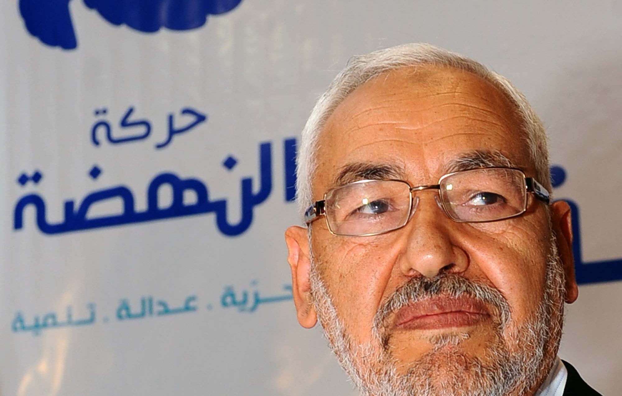 The image features a man with a gray beard and glasses, looking thoughtfully to the side. He is dressed in a light-colored shirt and blazer. In the background, there is a logo with Arabic text and a star, indicating it is related to a political movement or organization. The overall tone of the image suggests a serious or contemplative mood.