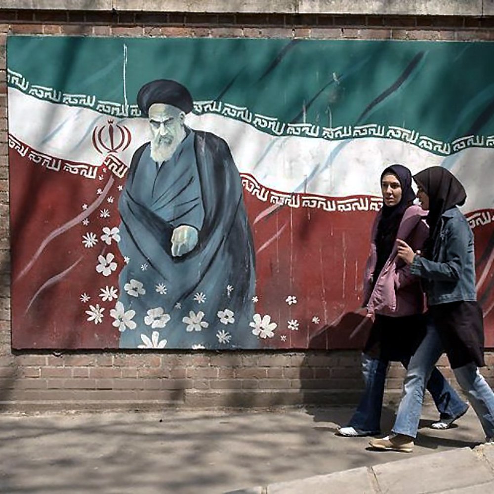 الصورة تظهر جدارية على جدار مبني، حيث تبرز شخصيتان متجولتان في الشارع. الجدارية تحتوي على رسم لقائد تاريخي إيراني، مع العلم الإيراني في الخلفية. الفتيات اللاتي يمررن ترتدين الحجاب، ويبدو أنهن في وضع محادثة. تتضمن الجدارية أيضاً بعض الزهور، مما يضفي لمسة جمالية على المشهد.