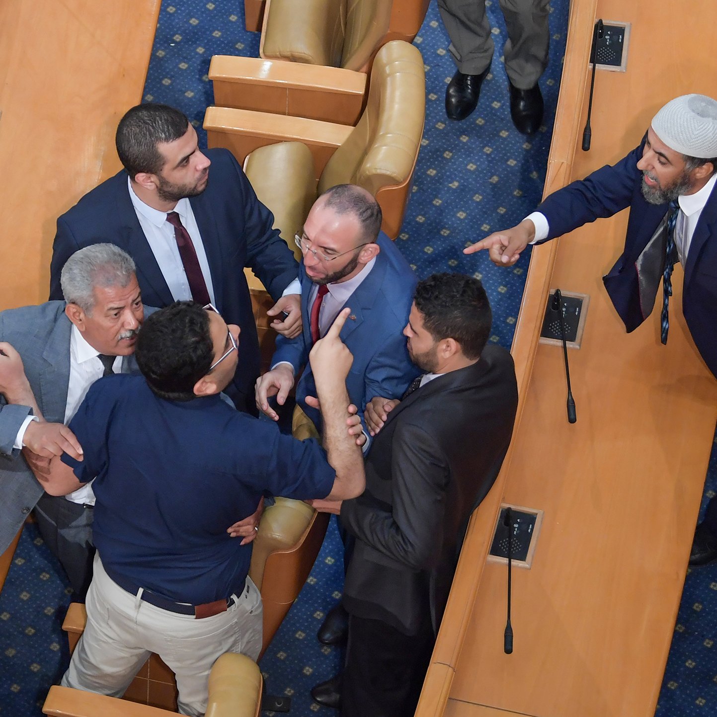 L'image montre une scène tendue dans une assemblée ou un parlement. Plusieurs hommes discutent vivement, certains pointant du doigt et affichant des expressions agitées. Il y a une mise en scène de désaccord, avec des gestes éloquents et une proximité physique entre les participants. Le décor est simple, avec des sièges en bois et un tapis bleu au sol, suggérant un environnement formel.