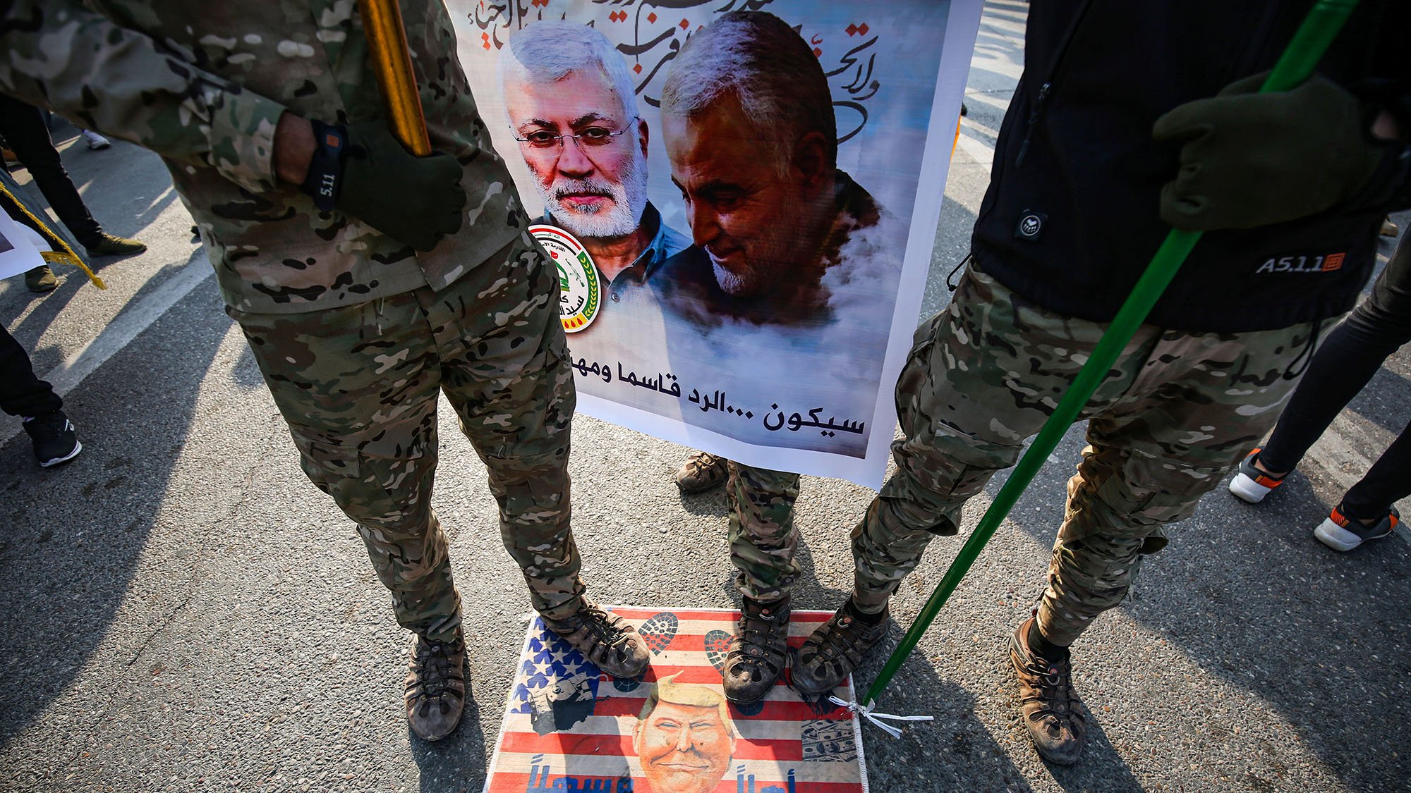 در این تصویر، تعدادی از افراد با لباس‌های نظامی و camo (رنگ‌های استتاری) در حال ایستادن هستند. آنها در دست خود عصا یا چوب‌هایی دارند. زیر پایشان تصویر یک پرچم آمریکاست که بر روی آن چهره‌ای در پایین مشاهده می‌شود. در پس‌زمینه، یک بنر بزرگ وجود دارد که شامل تصاویری از دو نفر است و متنی در مورد آنها نوشته شده است. فضایی حاکی از تظاهرات یا یک رویداد اجتماعی در حال شکل‌گیری است.